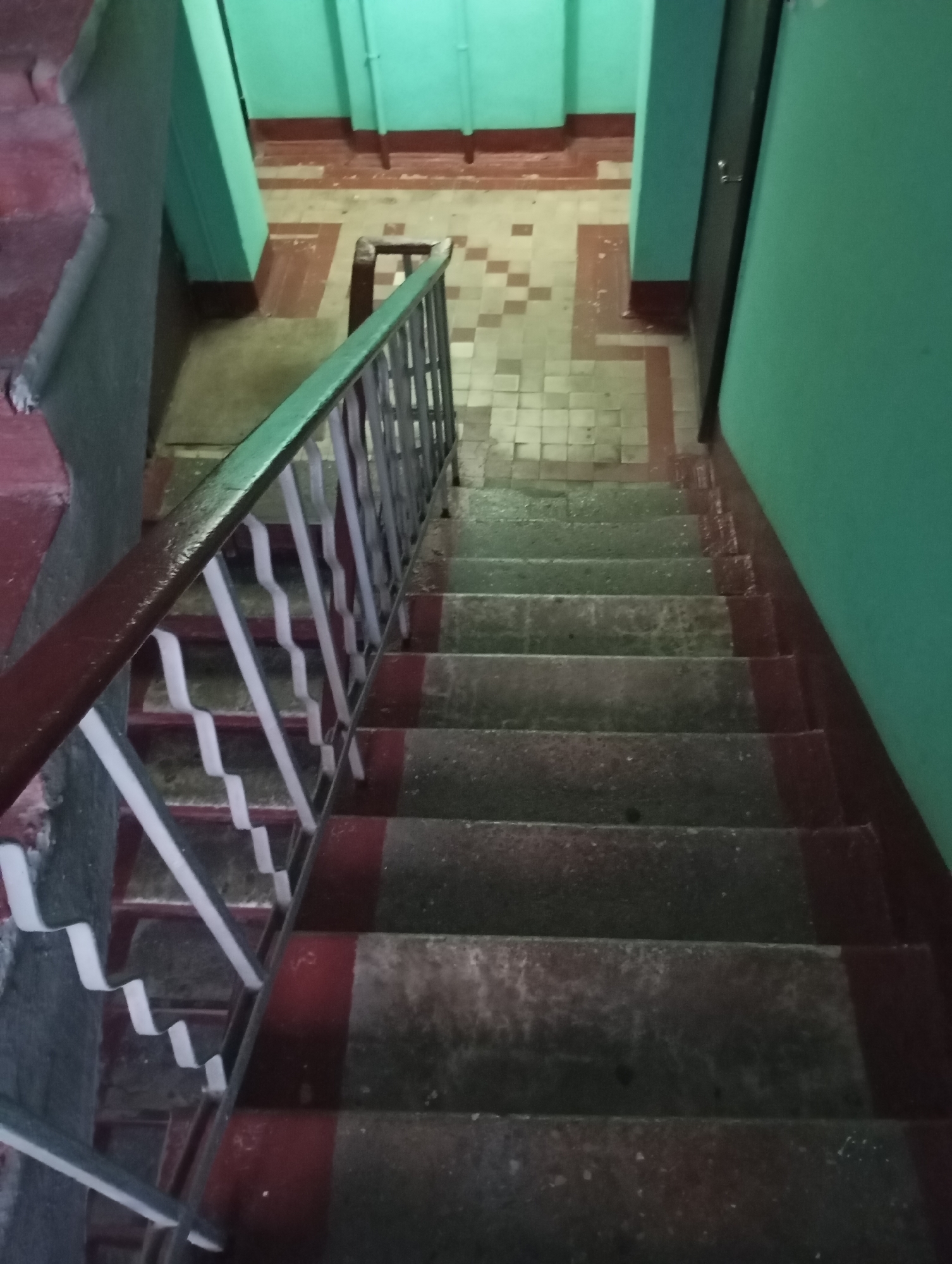 This is an interior photograph of a concrete staircase with red and dark gray painted risers and treads, leading downwards. A metal railing with a dark green handrail and white wavy supports runs along the left side of the stairs. The walls on the right are painted a light teal color. The floor at the bottom of the stairs is tiled with a pattern of light beige and dark red square tiles. The scene appears to be in a building, likely a residential building or an apartment complex, in Moscow, Russia. There are no people visible in the image, and no apparent actions or interactions taking place. The environment is dimly lit.