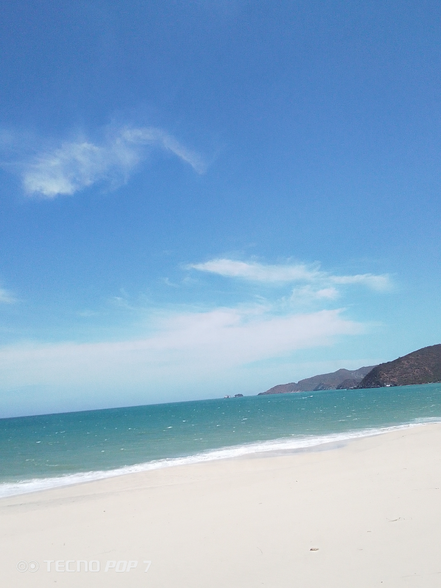 The image depicts a beach scene in Pedro González, Venezuela. The sky is blue with scattered clouds. The turquoise water meets a white sandy beach. Hills are visible in the distance. There are no people present. The image includes a watermark indicating it was taken with a TECNO POP 7 phone.