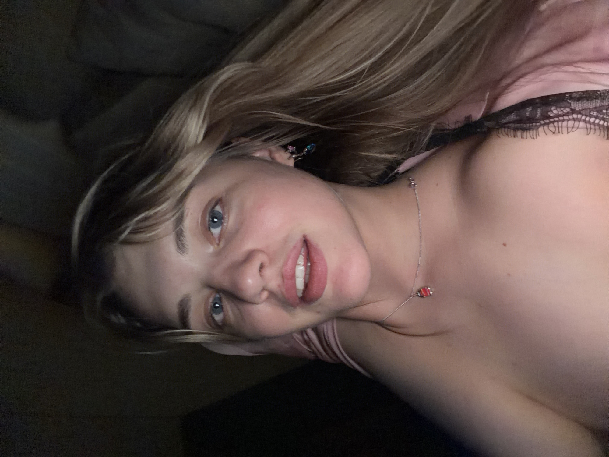A woman lies on a bed in a dimly lit room. She has blonde hair and blue eyes and is wearing a necklace with a small red pendant. The lace of her camisole is visible. Her earrings are visible. The location is unknown.