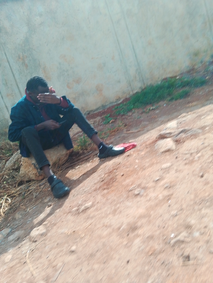 A young man, dressed in a dark blue jacket and black pants, is sitting on a rock. He is looking down, his left hand covering his face. He is wearing black shoes and a red shirt is visible underneath his jacket. There is a large rock behind him and the ground is dirt. He appears to be sitting on the edge of an unpaved road or path. There is short green grass in the background. This scene is located in Gitaru, Kenya, at coordinates -1.218, 36.655.