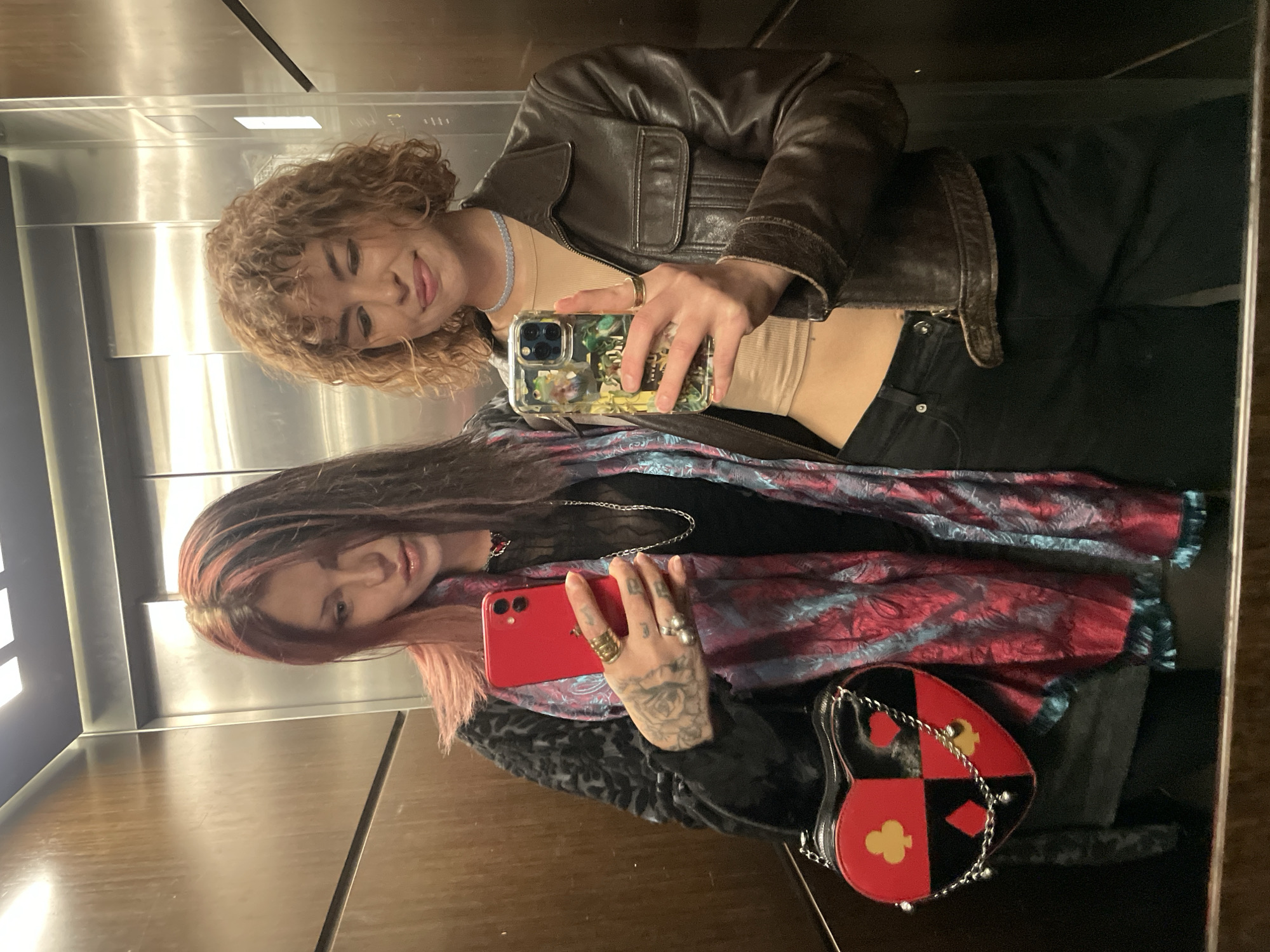 Two women are posing for a selfie in what appears to be an elevator. The woman on the top has curly brown hair, a brown leather jacket, a light-colored top, and dark pants. She is holding a phone with a decorative case. The woman on the bottom has long, dark hair with pink highlights and is wearing a dark top under a patterned scarf. She holds a red phone and has several rings and tattoos on her hand. There is a heart-shaped bag with card suit symbols visible. The elevator interior is composed of brushed metal and wood paneling. The location, Boreti, Montenegro, cannot be ascertained from the image.