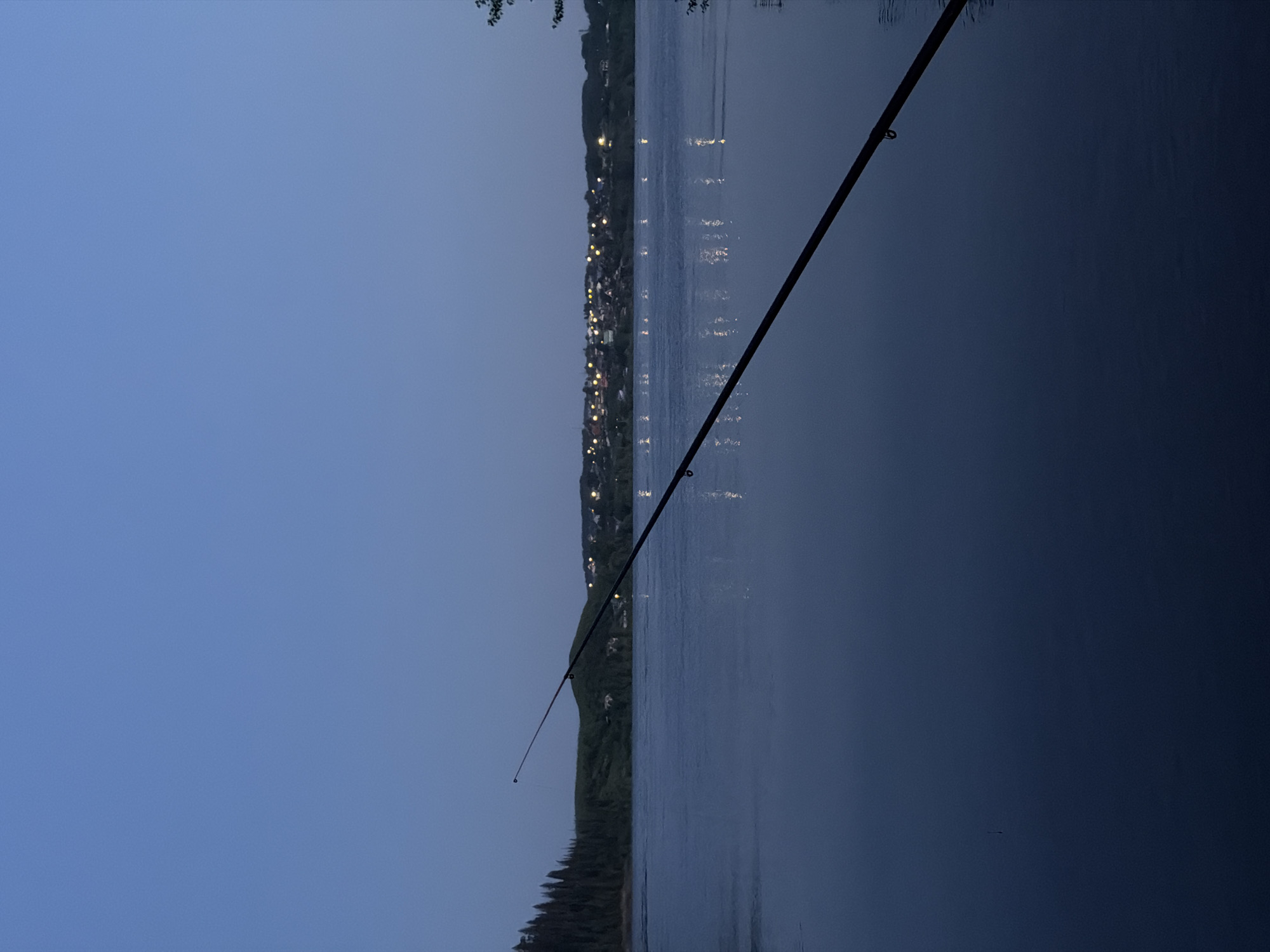 The image depicts a scene at dusk or night, possibly in the vicinity of Noviy, Russia, as inferred from visual cues resembling illuminated city lights along a body of water. A fishing rod is prominently positioned diagonally across the frame, suggesting someone is fishing. The water surface reflects some of the city lights, indicating calm conditions. A dark treeline or foliage is visible on the near side of the water. The sky is a gradient of blues, suggesting twilight or approaching darkness. No people are visible.