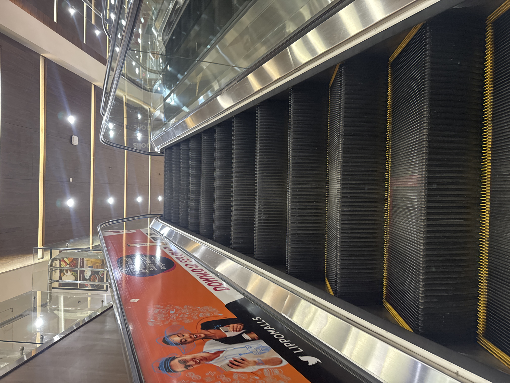 In Kuta, Indonesia, an escalator ascends in a shopping mall. The escalator features black steps with yellow safety lines. The left side of the escalator is adorned with an orange advertisement banner displaying illustrations of a man and a woman holding smartphones. The text on the banner includes "DOWNLOAD SEKARANG" and "LIPPOMALLS." The escalator is surrounded by polished metal railings and glass panels, reflecting the mall's interior lighting. Above, the mall ceiling is visible, featuring wooden paneling and integrated lighting fixtures.