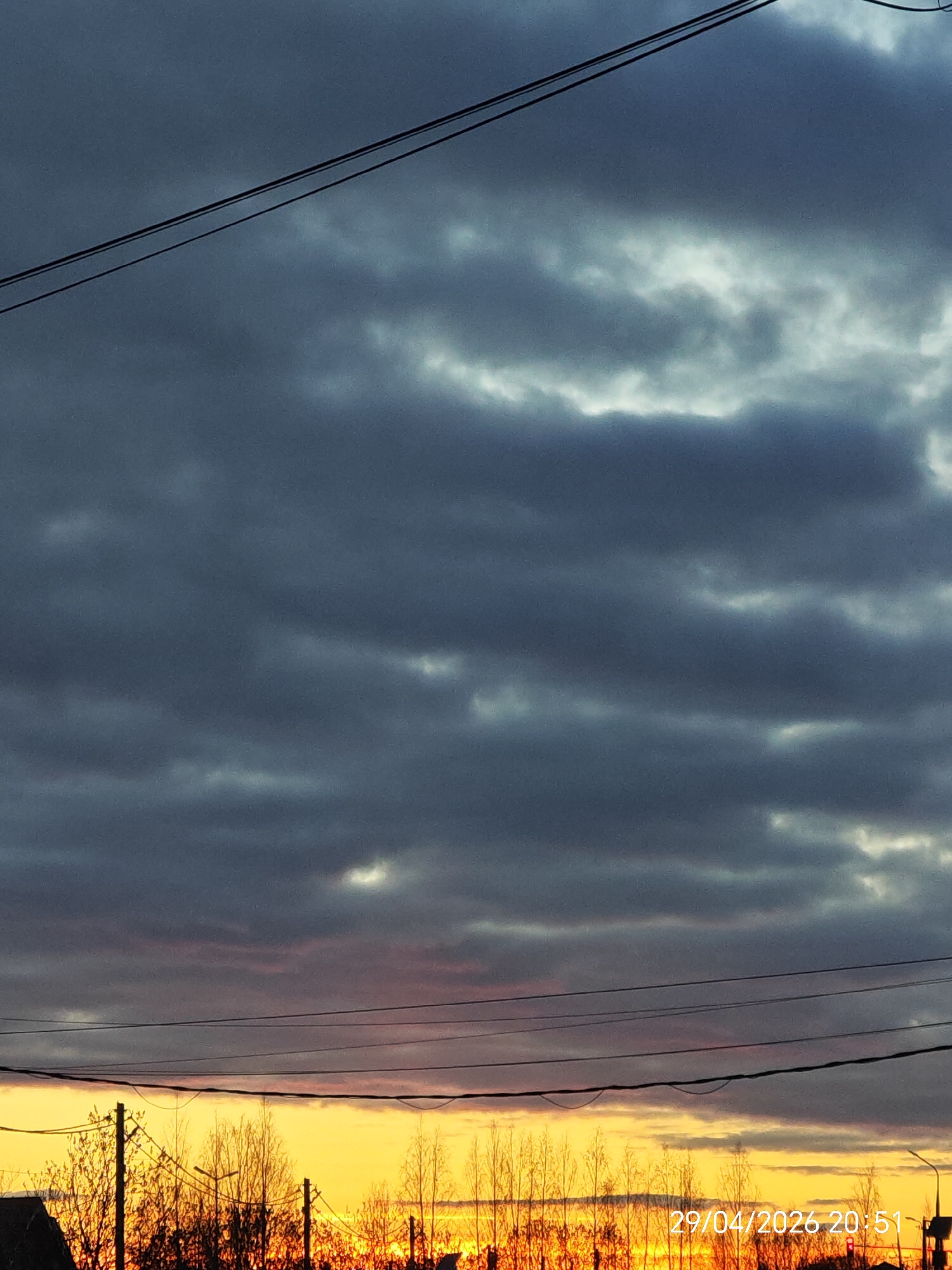 The image displays an outdoor scene at dusk, characterized by a dramatic sky. The upper portion of the frame is dominated by heavy, dark grey and bluish-grey clouds, with some lighter, brighter patches visible, suggesting varying cloud thickness or breaks in the overcast sky. Multiple dark power lines or utility wires stretch across the image horizontally, with one pair traversing the top and another set crossing the mid-section.
Below the cloud cover, the horizon glows with intense golden yellow and orange light, indicating a sunset or sunrise, with the sun itself hidden from view. Silhouetted against this bright glow are numerous bare trees or trees with sparse foliage, their dark branches forming intricate patterns. Several dark utility poles are also visible in silhouette among the trees, and hints of rooftops or other structures can be seen at the very bottom edges of the frame.
The overall setting appears to be a rural or semi-urban landscape. The time of day is confirmed by a digital timestamp "29/04/2026 20:51" in white text at the bottom right corner, indicating evening. No people are visible in the image. The weather is overcast but allows the vibrant colors of the setting sun to break through, creating a striking contrast with the dark clouds.