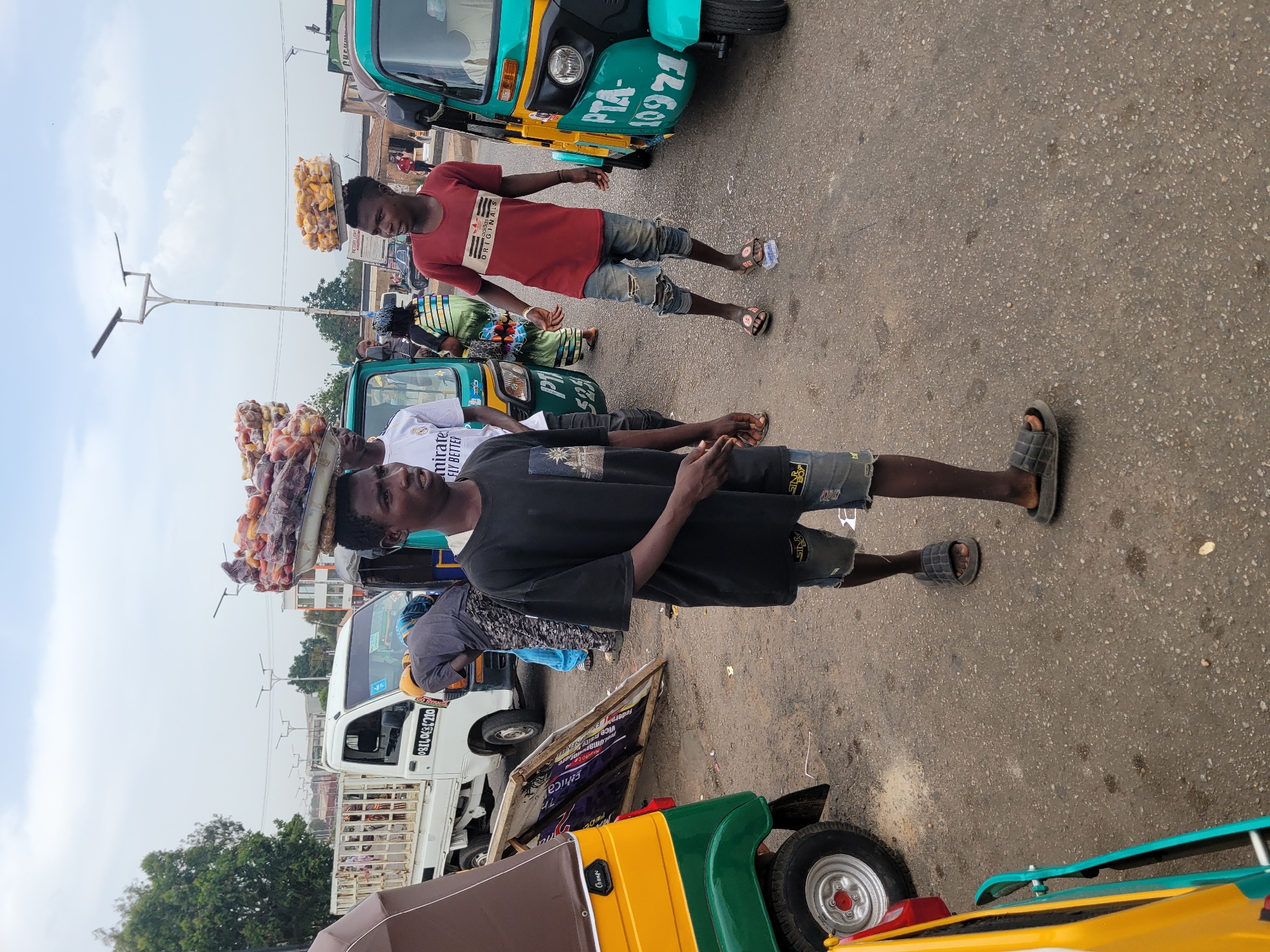 The image depicts a street scene in Jos, Nigeria. Two young men are walking on a paved road with various vehicles, including "Keke Napep" auto rickshaws and a white flatbed truck, parked nearby. One man carries a tray of what appear to be fruits or snacks on his head. Another man is carrying another tray. The vehicles have license plate and company advertisement. The scene appears to be a bustling commercial area.