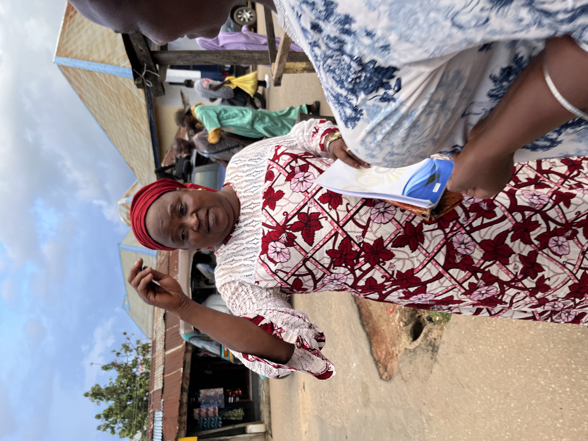 An outdoor scene in Jos, Nigeria, depicts a daytime interaction within what appears to be a bustling market or street. The main subject is a woman in the foreground, captured from the chest up, facing slightly towards the right side of the frame but looking directly at the viewer. She is wearing a vibrant white dress with a detailed pattern of red and pink flowers, green leaves, and geometric lines, complemented by white lace on the sleeves and neckline. A red headscarf is wrapped around her head. In her right hand, she holds several papers or a thin book with a colorful cover featuring blue and yellow elements. Her left hand is partially raised.
Partially visible in the upper right corner is another person, seen from the shoulder up, facing the main woman, suggesting a conversation or exchange. The background reveals a lively street environment with several blurred individuals and makeshift structures typical of an open-air market. These structures feature corrugated iron roofs, wooden supports, and canvas awnings. What appears to be a small shop with goods displayed on shelves can be seen in the distance. The ground is unpaved and dusty. The sky above is partly cloudy. No discernible text is clearly visible on the papers held by the woman or elsewhere in the scene.