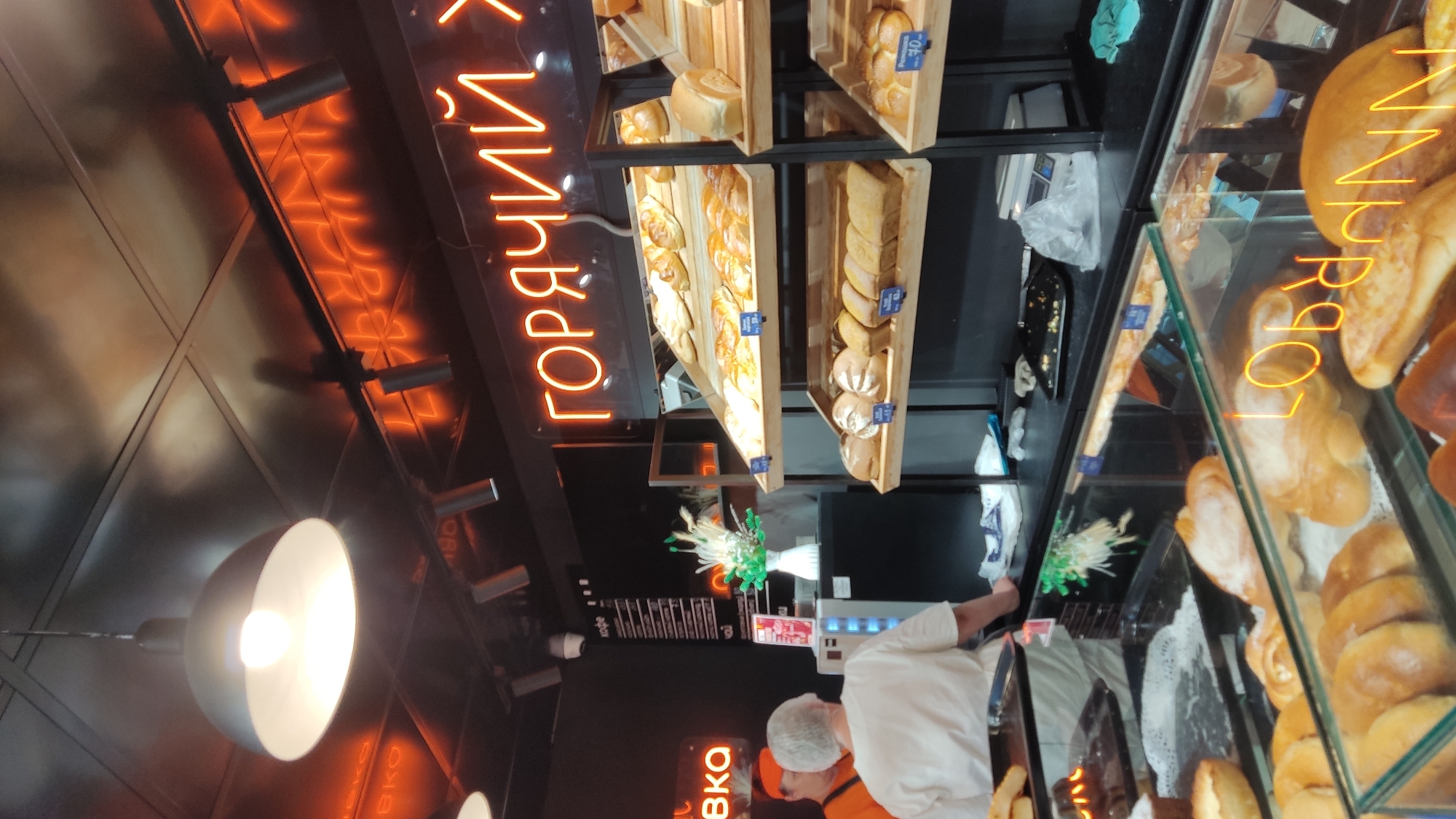 The image depicts the interior of a bakery, likely in Krasnodar, Russia, as indicated by the Russian text on the neon sign. The scene is dominated by baked goods displayed on wooden shelves and in a glass-enclosed counter.
In the background, a bright orange neon sign with Cyrillic lettering reads "ГОРЯЧИЙ", which translates to "HOT". The sign is mounted above a display shelf filled with various pastries and loaves of bread. Another smaller orange neon sign, partially visible on the left, reads "ВЫПЕЧКА", meaning "BAKERY PRODUCTS".
Two individuals are visible in the middle ground. One person, wearing an orange cap and shirt, appears to be a bakery employee. The other person, seen from the back and wearing a white shirt and hairnet, is likely a customer. They are standing near a counter where items such as coffee and tea are listed on a menu board mounted on the dark wall. A white vase with dried floral decorations sits on the counter between them.
In the foreground, a glass display case showcases a variety of bread and pastry items. The reflection on the glass reveals more of the bakery's interior, including overhead lighting and more neon signage. The overall ambiance suggests a bustling retail environment focused on fresh baked goods.