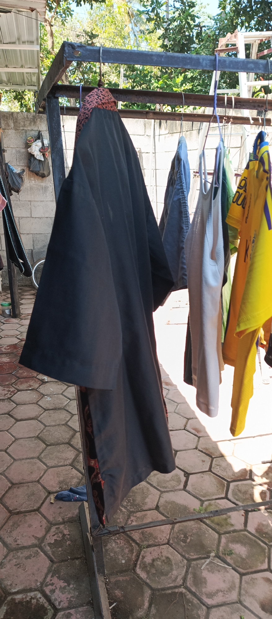 A black garment with a patterned trim is hanging on a clothesline. The garment is large and appears to be a robe or a dress. It is hanging alongside other items of clothing, including a blue shirt, a white tank top, and a yellow t-shirt. The clothesline is set up outdoors, with a brick wall and trees in the background. The ground is paved with hexagonal tiles.