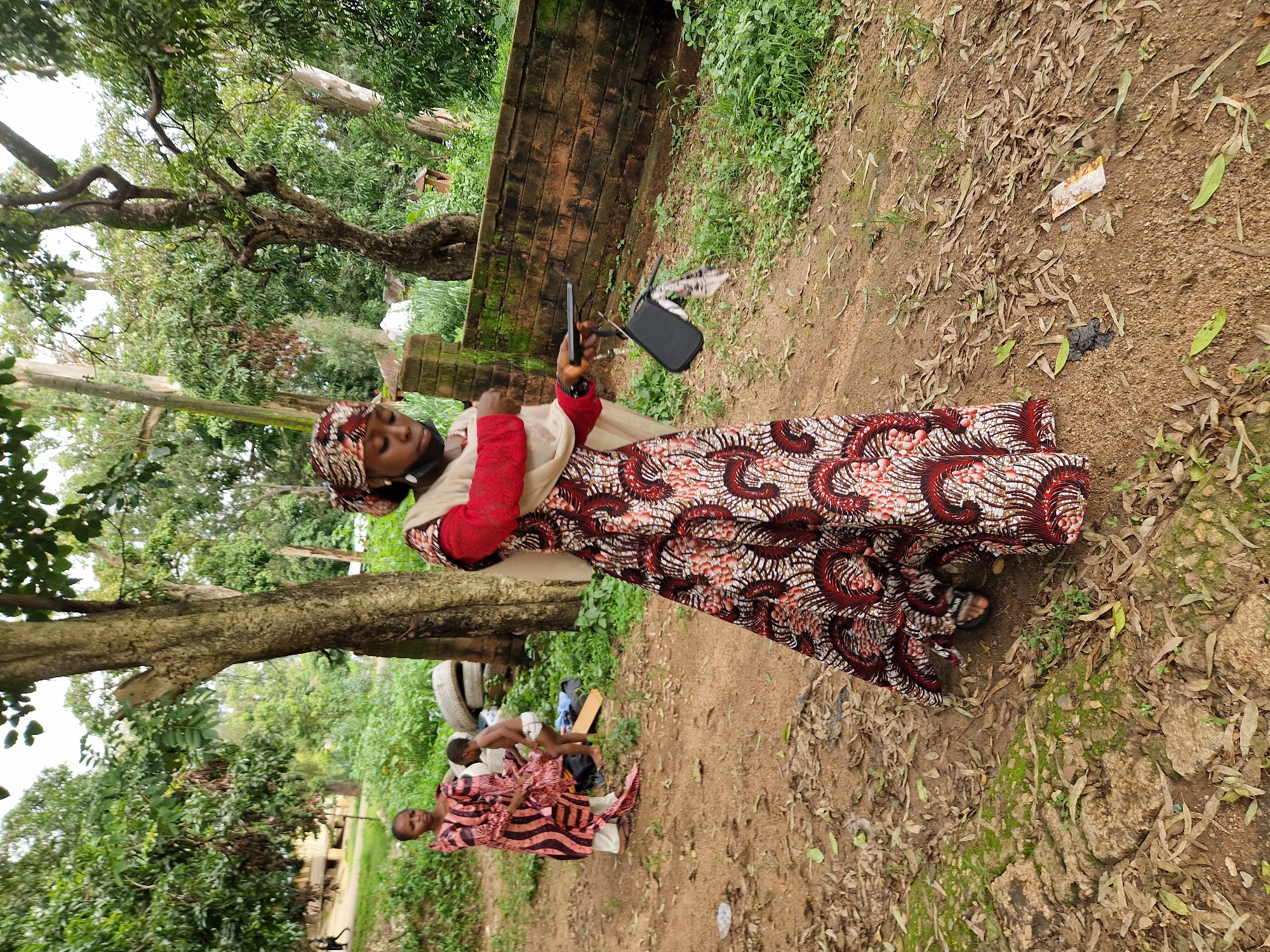 In Jos, Nigeria, an outdoor scene unfolds in what appears to be a somewhat neglected communal area. A woman, adorned in a vibrant, patterned African dress and headscarf, stands prominently in the foreground. Her dress features a striking red and white design with swirling motifs. She is wearing a light-colored shawl over a red long-sleeved top and a black face mask pulled down below her nose. In her right hand, she holds a dark-colored object, possibly a phone or a small purse, with a patterned fabric accessory attached to it.
Behind her, to the left, another woman in a similar patterned dress, though with a vertical stripe design, is interacting with a young child. The child, shirtless and wearing a diaper, is standing and seems to be adjusting clothing. In the background, several discarded tires are stacked.
The ground is covered in reddish-brown earth, scattered with dry leaves and some green vegetation. A low, weathered brick wall runs horizontally across the mid-ground. Trees with thick trunks and lush green foliage dominate the background, suggesting a possibly humid climate. The overall impression is one of everyday life in a community setting, with a focus on traditional attire and a natural, albeit somewhat unkempt, environment.