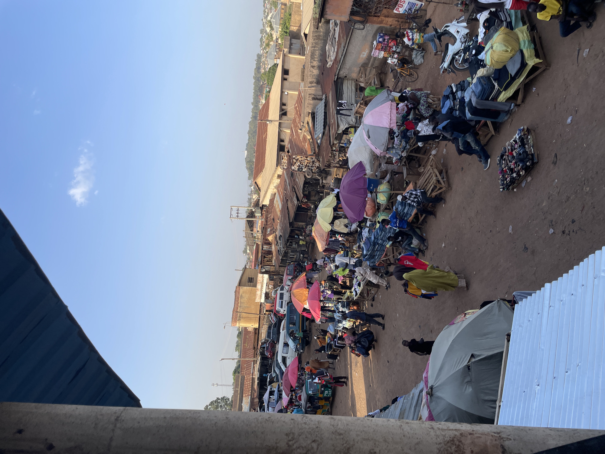 The image depicts an open-air marketplace in Bukuru, Nigeria, viewed from an elevated position. The scene is dominated by rows of stalls covered by umbrellas of various colors (pink, green, purple, and beige). Merchandise, primarily clothing, is displayed on wooden pallets and tables. People are scattered throughout the market, presumably shopping and selling. Vehicles, including vans and motorcycles, are parked along the side of the market. Buildings with corrugated metal roofs line the edges of the market area. The sky is clear and blue. The ground is reddish-brown dirt.