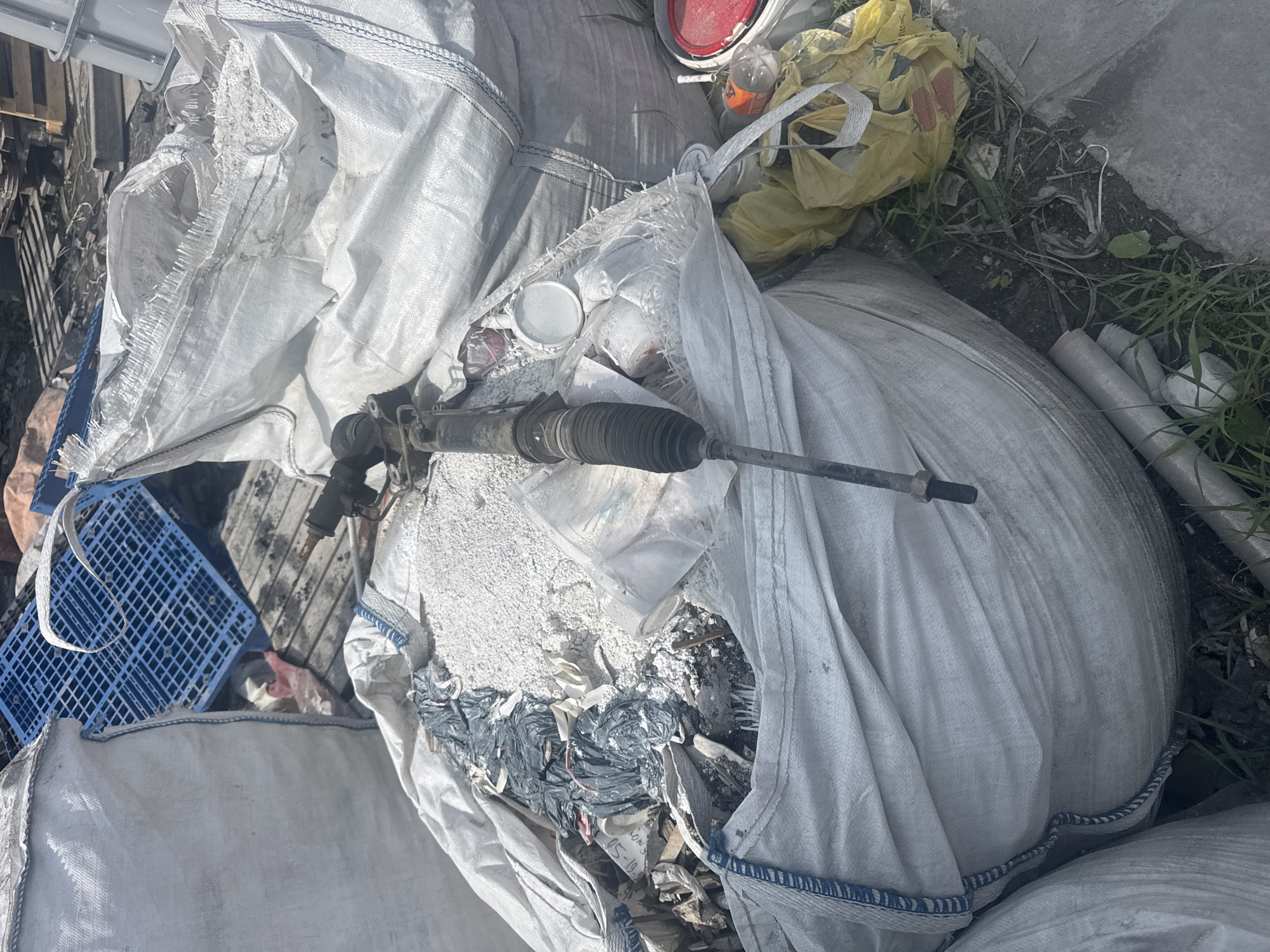 The image shows a close-up view of a pile of industrial waste. The primary focus is on a large white woven plastic bag, partially filled with white granular material and other debris. A black automotive steering rack component, possibly from a car or truck, is visible protruding from the bag. The steering rack has a ribbed rubber boot and metal components. Other items in the frame include a blue plastic grid, various smaller bags (one is yellow), a red-lidded bucket, and scattered debris on the ground which appears to be dirt, grass, and miscellaneous waste materials. The scene is outdoors, likely in a waste disposal or construction site area. The presence of industrial-sized bags and discarded automotive parts suggests a context of bulk waste management or recycling. The lighting indicates it is daytime. No people are visible in the image. The location is identified as Metallostroy, Russia.