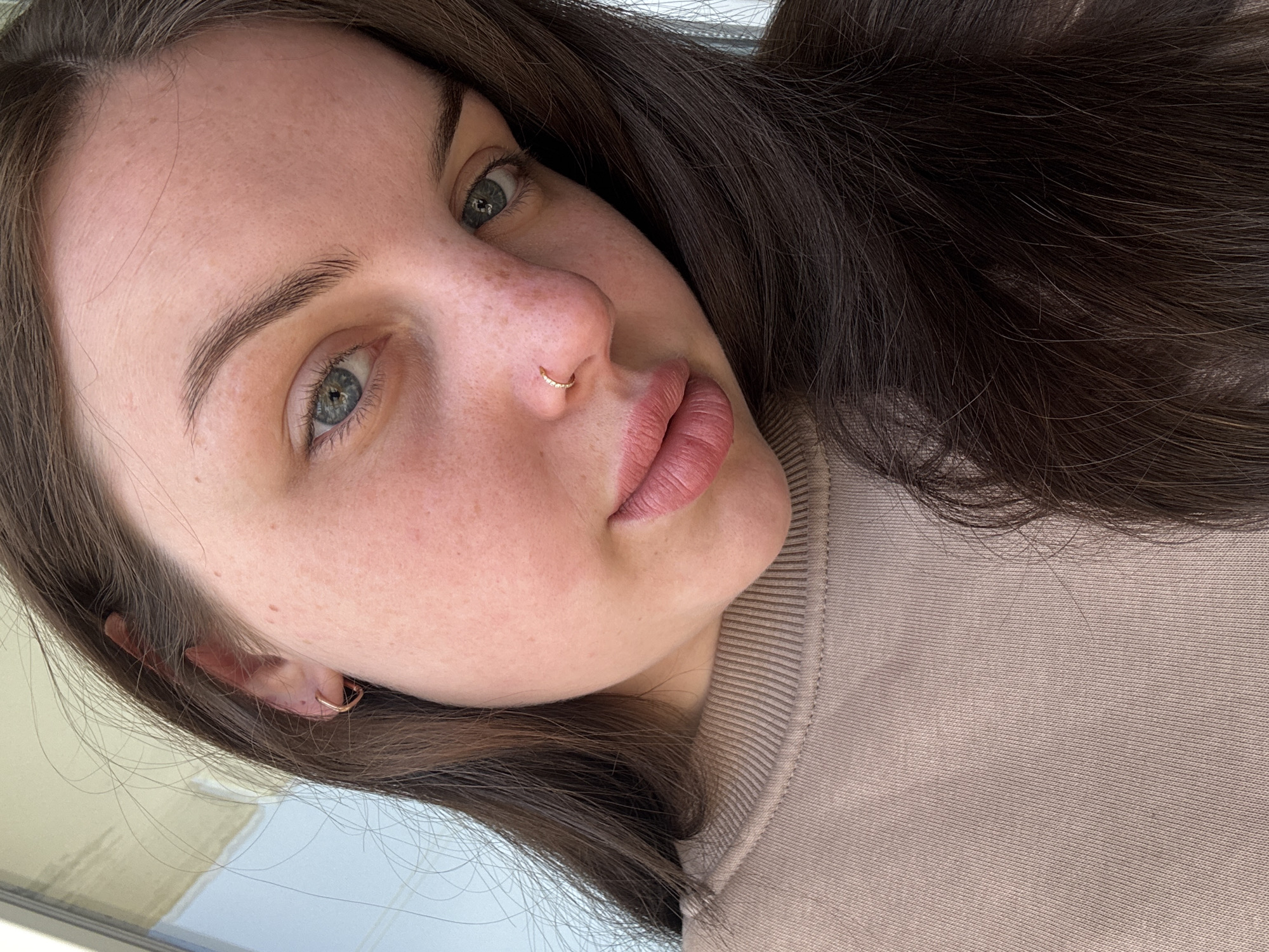 A close-up, slightly rotated portrait captures a young woman from the chest up, with a primary focus on her face. She has long, dark brown hair that frames her face, blue-green eyes, and natural-looking, defined eyebrows. Her fair skin shows subtle freckles across her forehead, cheeks, and nose. A small, golden hoop nose ring is visible on her left nostril, and a matching golden hoop earring adorns her left earlobe. Her lips are full and have a matte, pinkish-nude color. She maintains a neutral to slightly pouting expression, gazing directly towards the viewer. She is wearing a light brown or beige crewneck sweatshirt with a ribbed texture, visible around her neck and shoulder. The lighting is soft and even, suggesting an indoor setting, possibly near a window, with an indistinct, light-colored background. No text is visible in the image. The scene is located in Chornomorskoe, Ukraine.
