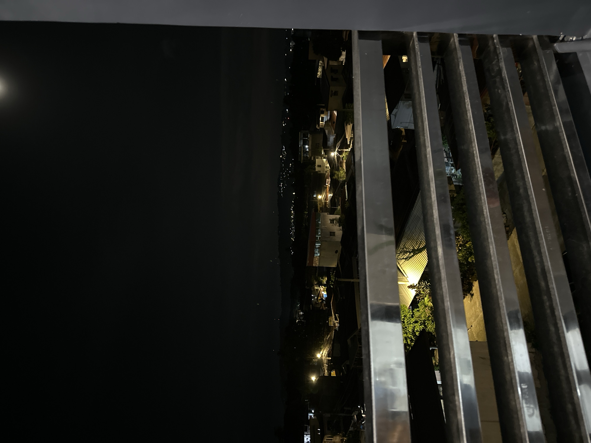 A night scene is captured from a high vantage point, possibly a balcony or elevated platform, in Davao City, Philippines. A metal railing runs diagonally across the right half of the frame. Beyond the railing, city lights twinkle, interspersed with buildings and vegetation. The left half of the frame shows a dark body of water under a night sky with a visible moon.