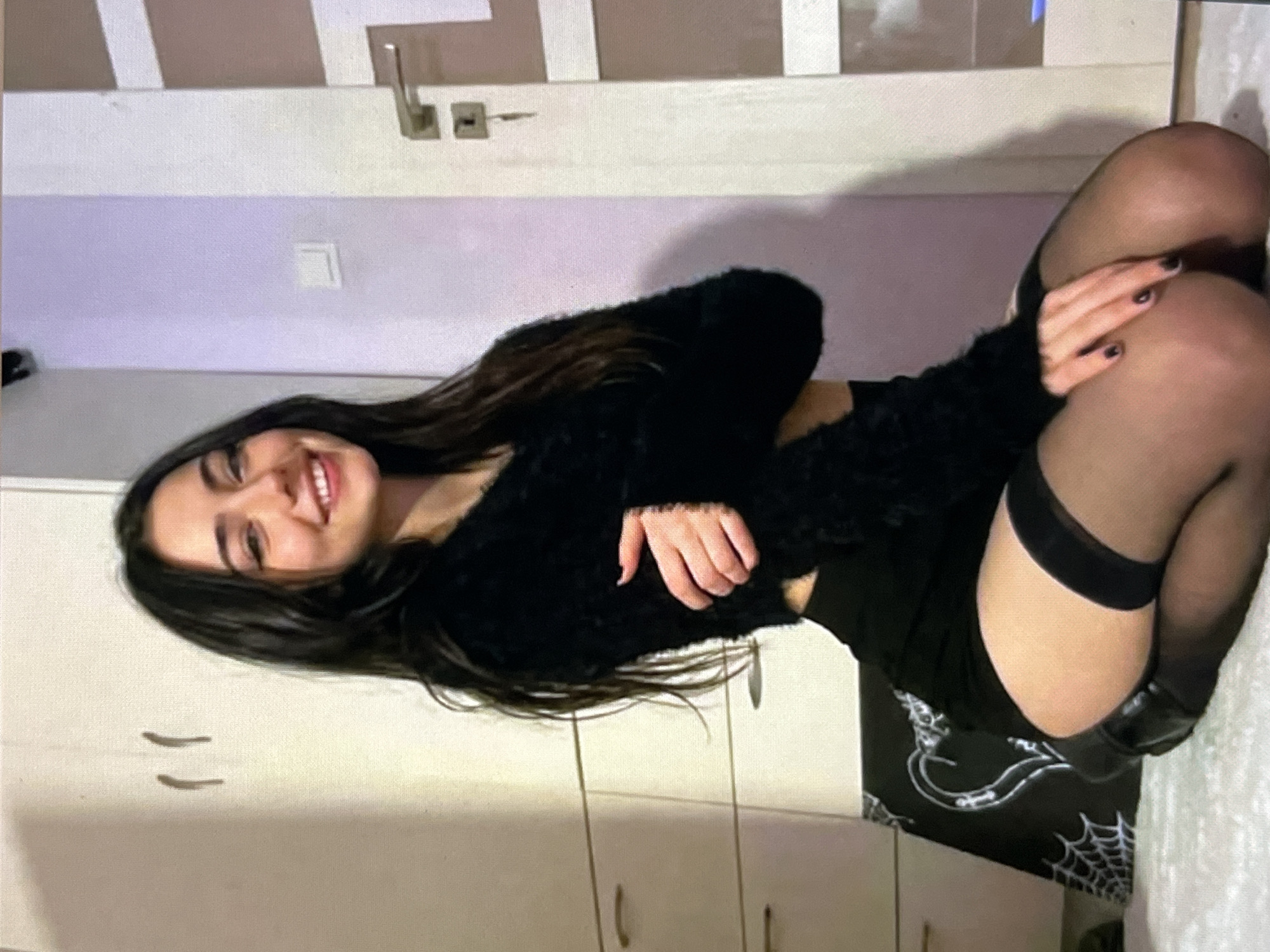 A young woman with long, dark hair and fair skin is kneeling on a light-colored rug in an indoor setting. She is wearing a black, fluffy, cropped sweater, a black mini-skirt, and black thigh-high stockings. Her arms are crossed in front of her. She is smiling and looking towards the camera. Behind her, to the left, is a white wardrobe with curved handles. To the right, a door with a silver handle and a keyhole is visible. The wall behind her is painted a light purple color. On the floor to her left, there is a black object with white spiderweb and heart designs.