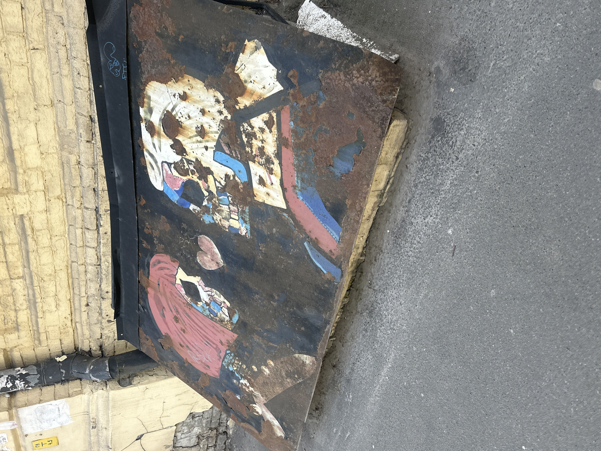 The image shows a rusty, metal door or panel leaning against a yellow brick wall and partially resting on the sidewalk. The panel has a faded, graffitied design depicting figures and shapes, including what appears to be a skull. Rust and peeling paint are evident across the surface. The sidewalk is gray and textured. The location is Kyiv, Ukraine.