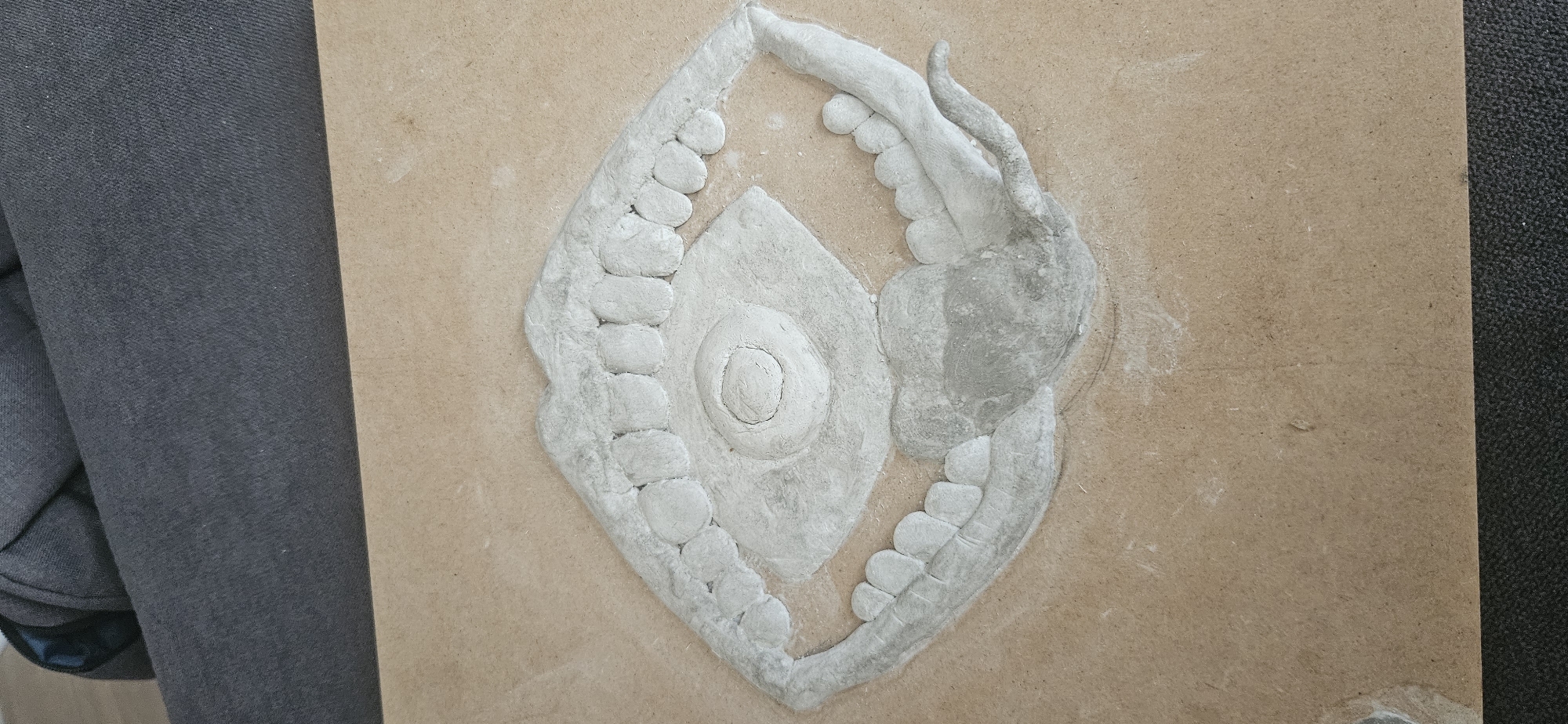 The image shows a partially completed bas-relief sculpture on a flat, light-brown surface, likely a piece of wood or particle board. The sculpture is symmetrical around a central vertical axis and consists of a diamond-shaped central element with a raised circular form in the center. Surrounding the diamond shape on either side are curving, arm-like appendages outlined with rounded beads. One arm appears to have additional sculptural details, possibly depicting fingers or claws. The sculpture is made of a pale, cement-like material. The sculpture is incomplete and shows areas of unfinished construction. To the left, a dark gray upholstered item, possibly a chair or sofa, is visible. To the right, a dark, slightly textured material, perhaps carpet, also appears. The background shows no indication of the location of the sculpture in the city of Plovdiv, Bulgaria.