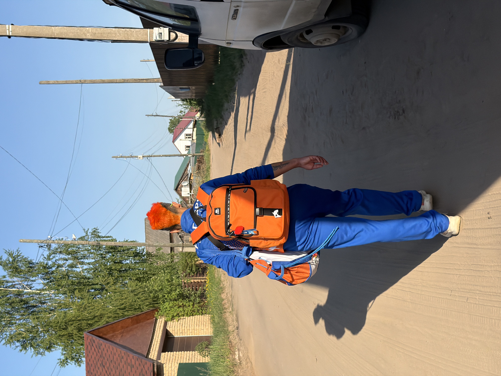 The image shows a person walking along a sandy road in a residential area. The person is wearing a blue tracksuit, white sneakers, and an orange backpack. The person's hair is bright orange. A white van is parked at the top of the frame. Houses and trees line the road. Power lines are visible above. The location is Chaikovsky, Russia.
