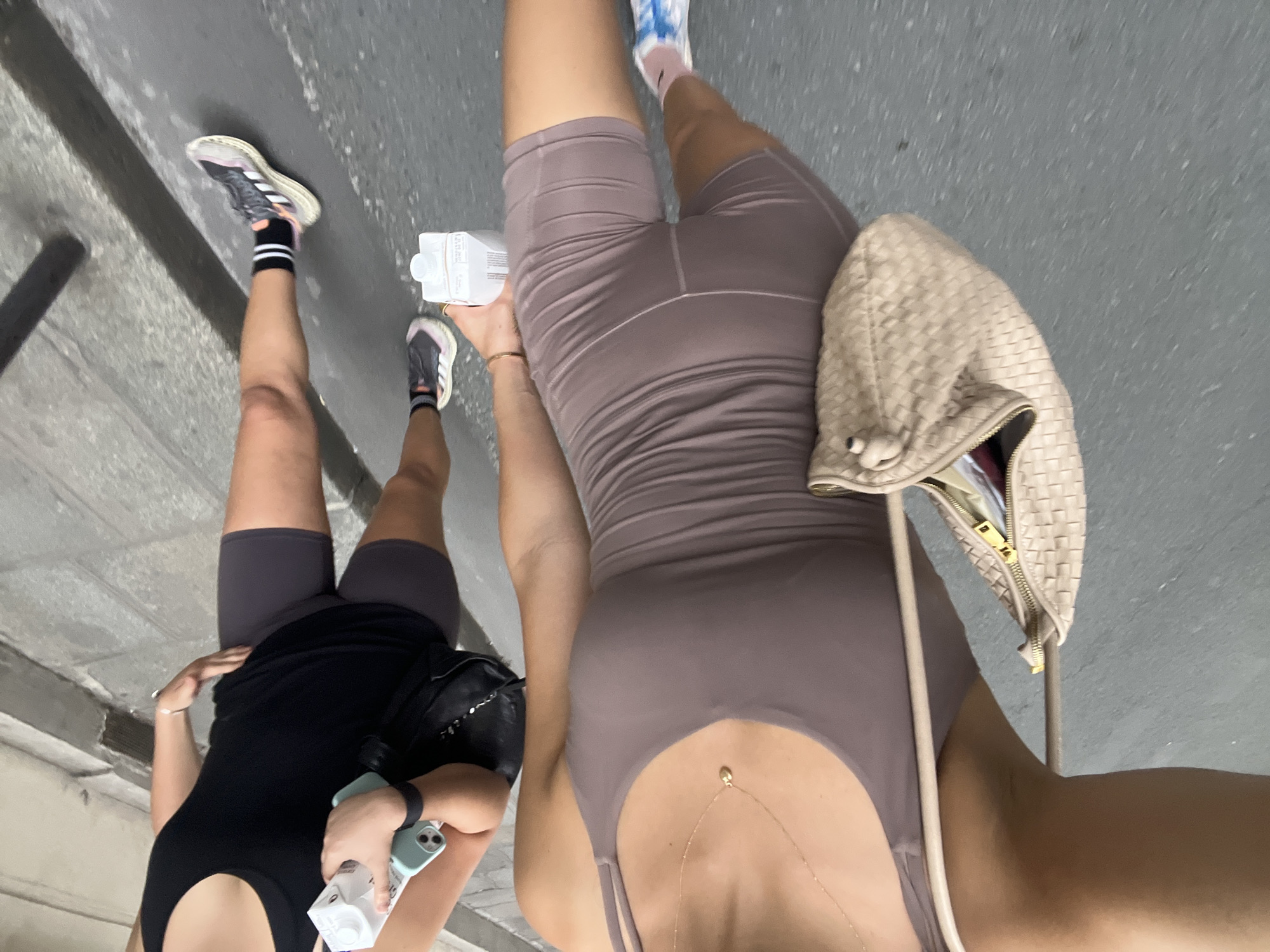 The media file shows two people, likely women, walking outdoors on a paved surface. The perspective is from above and slightly behind them, creating a sense of motion as they walk away from the camera. The lighting suggests it is daytime, possibly overcast, as there are no harsh shadows.
The person in the foreground is wearing a mauve-colored, form-fitting athletic outfit, including shorts and a tank top. They are carrying a beige woven handbag over their shoulder and a white carton of liquid in their right hand. A delicate gold necklace is visible on their chest.
The person in the background is wearing a black tank top and dark gray athletic shorts. They are holding a light blue smartphone and a white carton of liquid. Both individuals are wearing athletic shoes and socks.
The background appears to be an urban environment with a gray, textured ground surface that could be a sidewalk or pavement. There are some vertical elements in the background, possibly walls or structures, that are out of focus. One visible detail on the white carton reads "SPARKLING WATER".
The overall impression is of a casual outing, possibly a walk or exercise session. The scene is unremarkable and does not provide specific clues about the exact location within Paris, France, beyond the provided context. There are no strong emotional cues visible, but the activity suggests a leisurely or active pursuit.