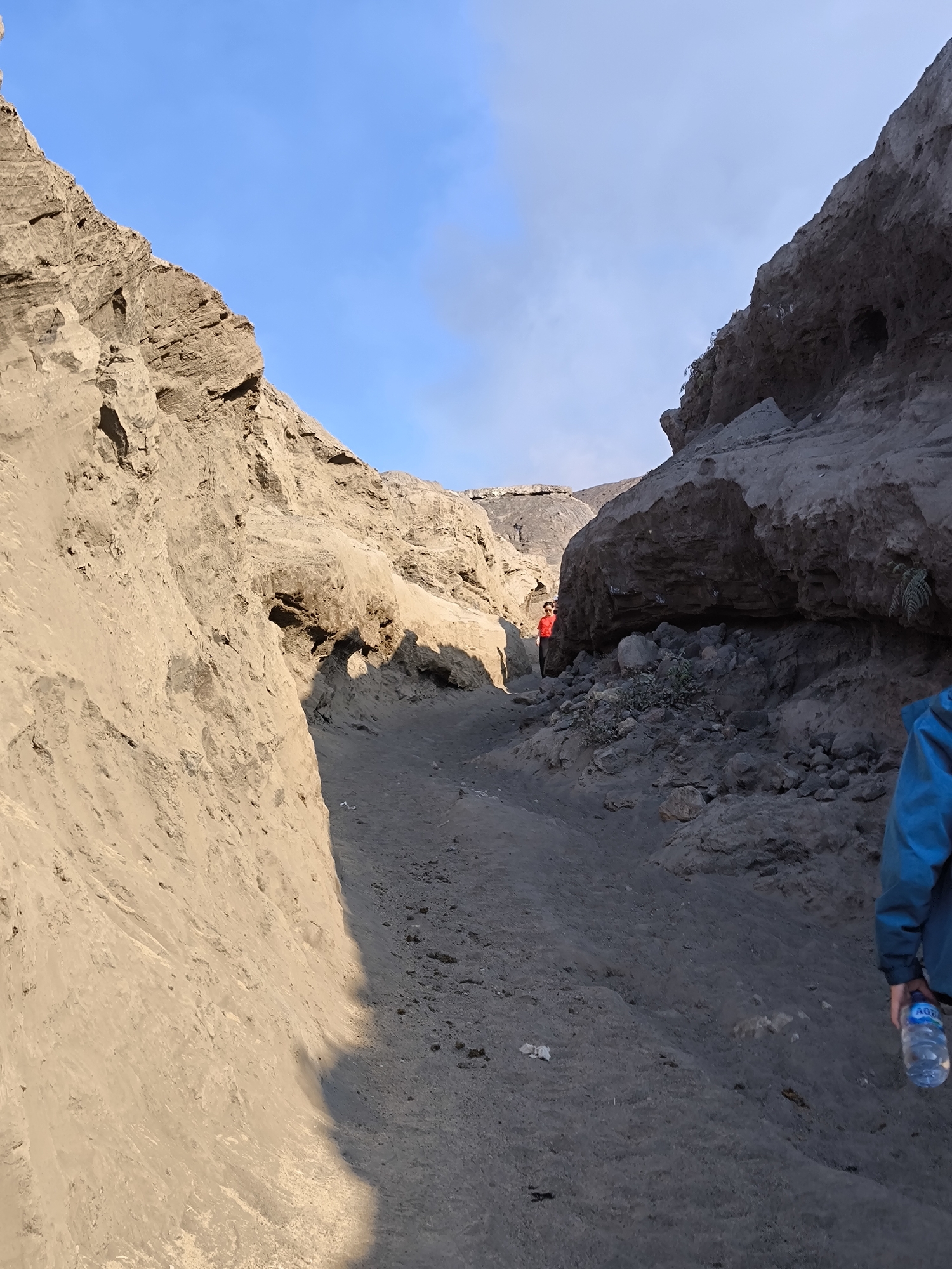 The image depicts a rocky, sandy terrain with two individuals. One person, wearing a blue jacket and holding a water bottle, is partially visible on the right side of the frame, walking away from the camera. Further into the scene, another person in a red shirt is walking up a sandy path between two large rock formations. The path appears to be part of a volcanic landscape, possibly Bromo Tengger Semeru National Park, given the location context of Cemorolawang, Indonesia. The sky is a clear blue, suggesting it is daytime, likely morning or afternoon due to the presence of shadows. The overall scene conveys a sense of outdoor adventure or hiking in a rugged environment. The water bottle is labeled with "AQUA."