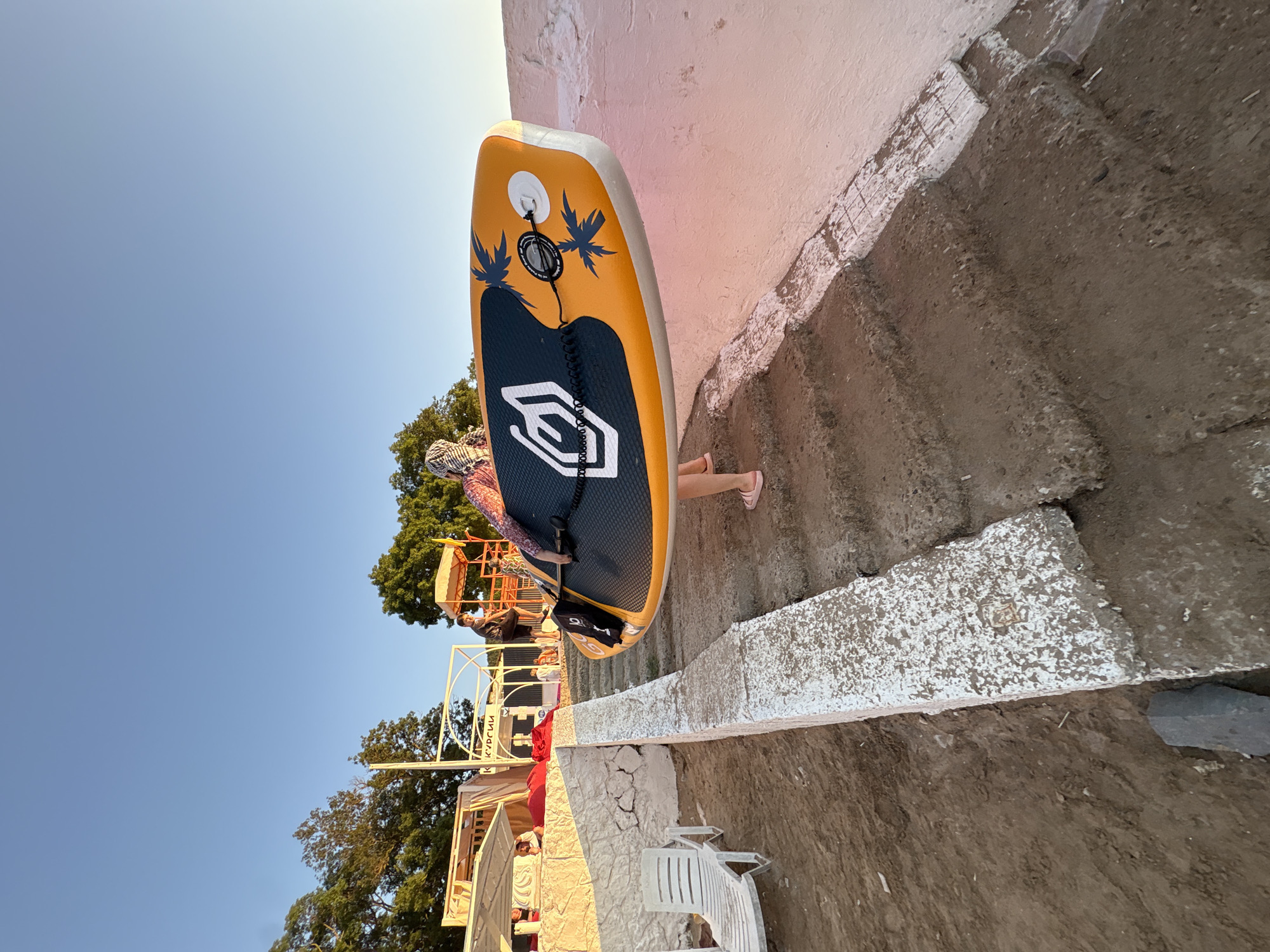 A person walks up a concrete staircase on a beach, carrying a paddleboard. The paddleboard is orange and black with a white logo and palm tree graphics. The person is wearing a headscarf and pink sandals. The background shows trees, a beach chair, and a structure that appears to be a beach cafe. The sky is clear and blue, suggesting it is daytime. The scene is likely located in the city of Novorossiysk, Russia, given the context of a beach environment.