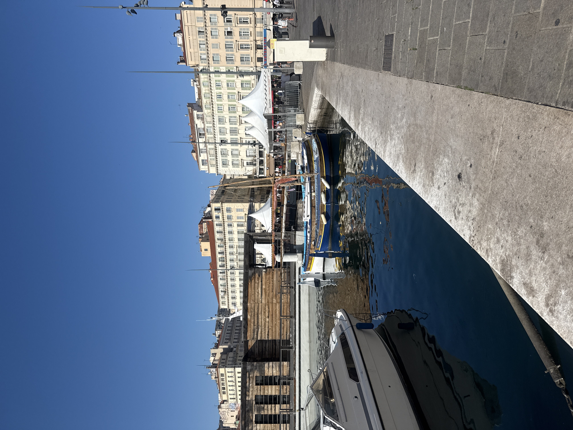 The image shows a waterfront scene in a city. In the foreground, a white speedboat is docked on the left, its reflection visible in the dark blue water. To the right of the speedboat, a traditional blue and yellow wooden boat with a mast is also docked. The water is calm, reflecting the boats and the sky.
Behind the boats, there is a stone quay with a paved walkway. Further back, a low, wooden building with horizontal planks and dark rectangular windows stands on the left. Beyond this building, several white tents are set up, suggesting a market or an event.
In the background, a row of multi-story buildings with pale facades and red tile roofs lines the waterfront. The architecture appears European. The sky is a clear, bright blue.
A few people are visible in the mid-ground, near the tents and walkways, some appearing to be walking or standing. The overall scene suggests a sunny day in a harbor area. The location is identified as Unknown, Unknown.