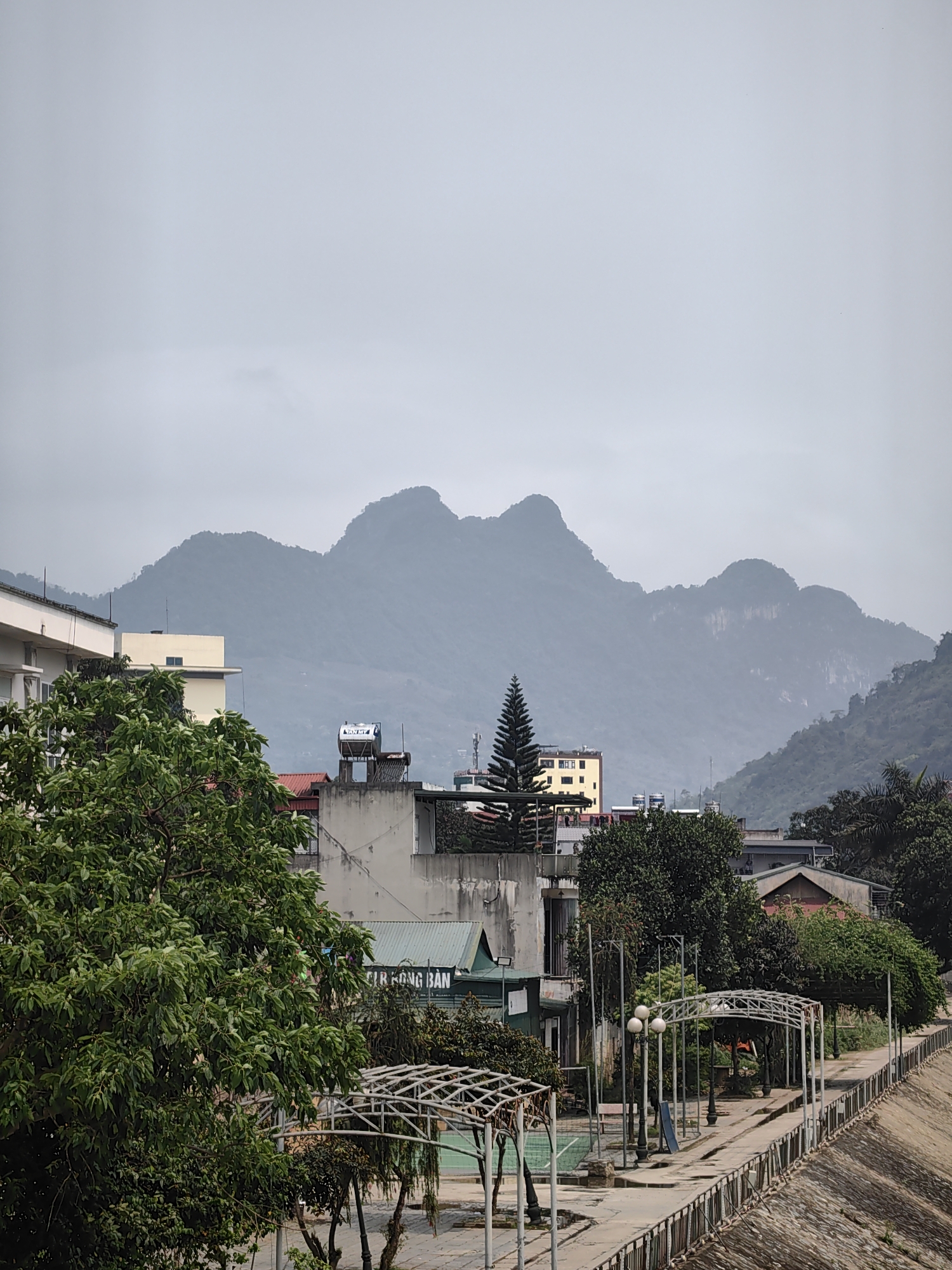 The photo shows a cityscape in Hà Giang, Vietnam, under an overcast sky. In the foreground is a park with metal arches, benches, and street lamps along a concrete pathway with a railing. Trees with green foliage are scattered throughout the scene. Behind the park are buildings of varying heights and architectural styles, including a light-colored building, a building with a water tank on the roof, and a building with a conical evergreen tree in front. The background features a mountain range obscured by mist. The overall environment appears damp and urban with the mountains providing a natural backdrop.