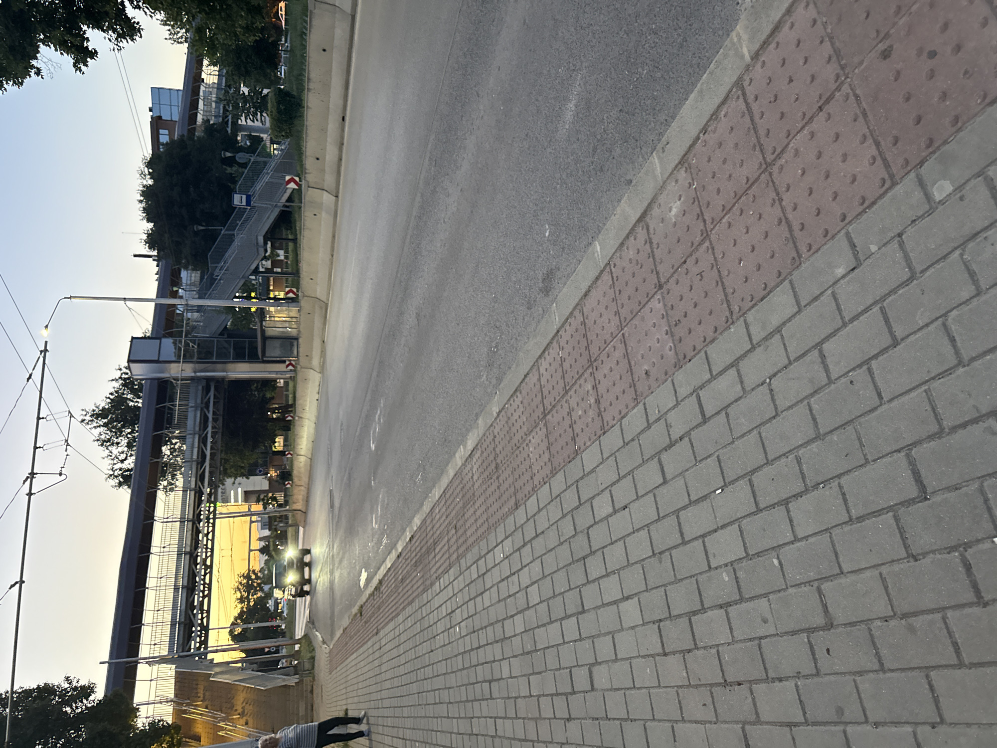 A person stands on a pedestrian walkway made of grey and red textured paving stones next to a road. The walkway has a tactile paving surface made of red tiles with raised dots, indicating a potential hazard or a crossing. The road is asphalt and appears to be a multi-lane street. In the background, there is a pedestrian overpass with stairs and ramps, likely crossing the road. A car with its headlights on is driving on the road under the overpass. The sky is a soft gradient of orange and blue, suggesting either sunrise or sunset. Trees and buildings are visible in the distance. Overhead power lines are also present.