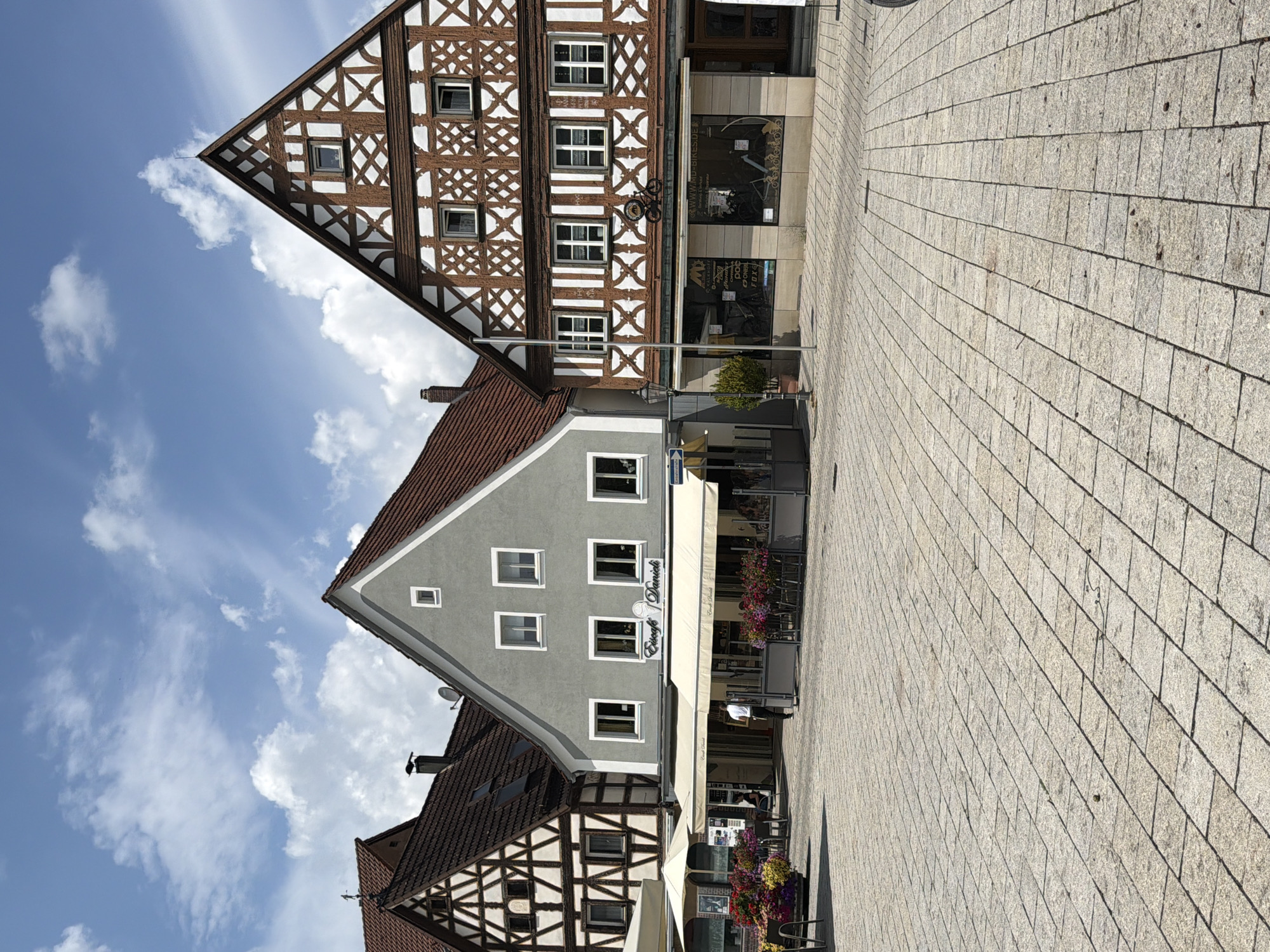 The image depicts a sunny day in Ebermannstadt, Germany, in front of a row of buildings. The foreground is a paved square with large, light-colored stone tiles. In the middle ground, there are three buildings. The leftmost building is a timber-framed house with a dark tiled roof and decorative white wooden beams. To its right is a grey-colored building with white-framed windows and a cream-colored awning extending over the sidewalk. The sign above the awning reads "Eiscafé Daniela." In front of this cafe, there are outdoor tables and chairs, some with umbrellas, and numerous pots of colorful flowers. A person in a white shirt is walking away from the viewer towards the cafe. The rightmost building is also a timber-framed structure with a similar decorative style to the leftmost one, featuring white beams on a dark wood background. This building has large display windows on the ground floor, one of which shows bicycles and related merchandise, suggesting it is a bike shop. A metal silhouette of a bicycle is mounted on the facade above the shop windows. The sky is blue with scattered white clouds.