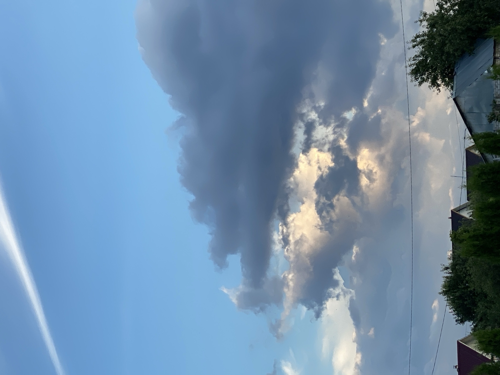 The image depicts a cloudy sky over Oktyabr'skii, Russia. The scene is dominated by a large, dark cloud formation in the center, with patches of lighter, illuminated clouds scattered around it. The sky is mostly blue, suggesting it might be late afternoon or early evening, as indicated by the interplay of light and shadow on the clouds.
In the right portion of the image, parts of buildings and trees are visible. The buildings have dark, sloped roofs, and one appears to be made of brick. Dense green foliage from trees surrounds the structures, hinting at a suburban or rural setting. A thin, white contrail is visible in the upper left quadrant of the image, suggesting an airplane passed overhead recently.
There are no people or animals visible in the image. The overall mood is somewhat dramatic due to the heavy cloud cover, but the presence of the blue sky and illuminated clouds prevents it from appearing entirely somber. No text is visible in the image.