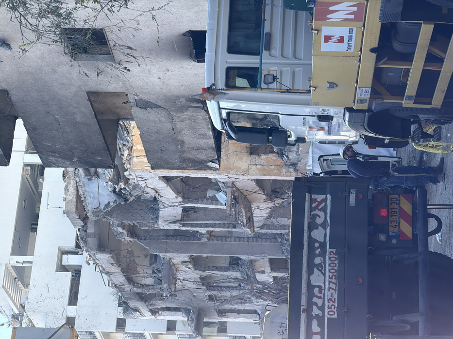 A daytime street-level image depicts a scene of severe structural damage and active work in Tel-Aviv, Israel. In the background, a multi-story building exhibits extensive collapse across its lower and middle floors, with shattered concrete and exposed rebar indicating significant structural failure. To the immediate right of the damaged building, a adjacent structure shows a weathered facade with darker, possibly charred, sections and visible window openings.
In the foreground, two large commercial vehicles are present. On the left, a dark-colored dump truck is viewed from the rear, displaying an Israeli license plate numbered "43-191-30" and Hebrew text with the phone number "052-7750002" on its tailgate. To its right, a white utility truck, partially obscured, is identified by the "NM" logo and Hebrew text "חברת חשמל לישראל" (Israel Electric Corporation) on its side. A small white flag with an orange stripe is attached to the utility truck's front corner. Between these two vehicles, a single male worker, wearing a white hard hat, dark blue long-sleeved shirt, blue jeans, and work boots, is crouched and focused on an area near the front wheel of the white utility truck. He appears to be holding a tool, possibly inspecting or working on the vehicle or the ground immediately beneath it. The sky above is clear.