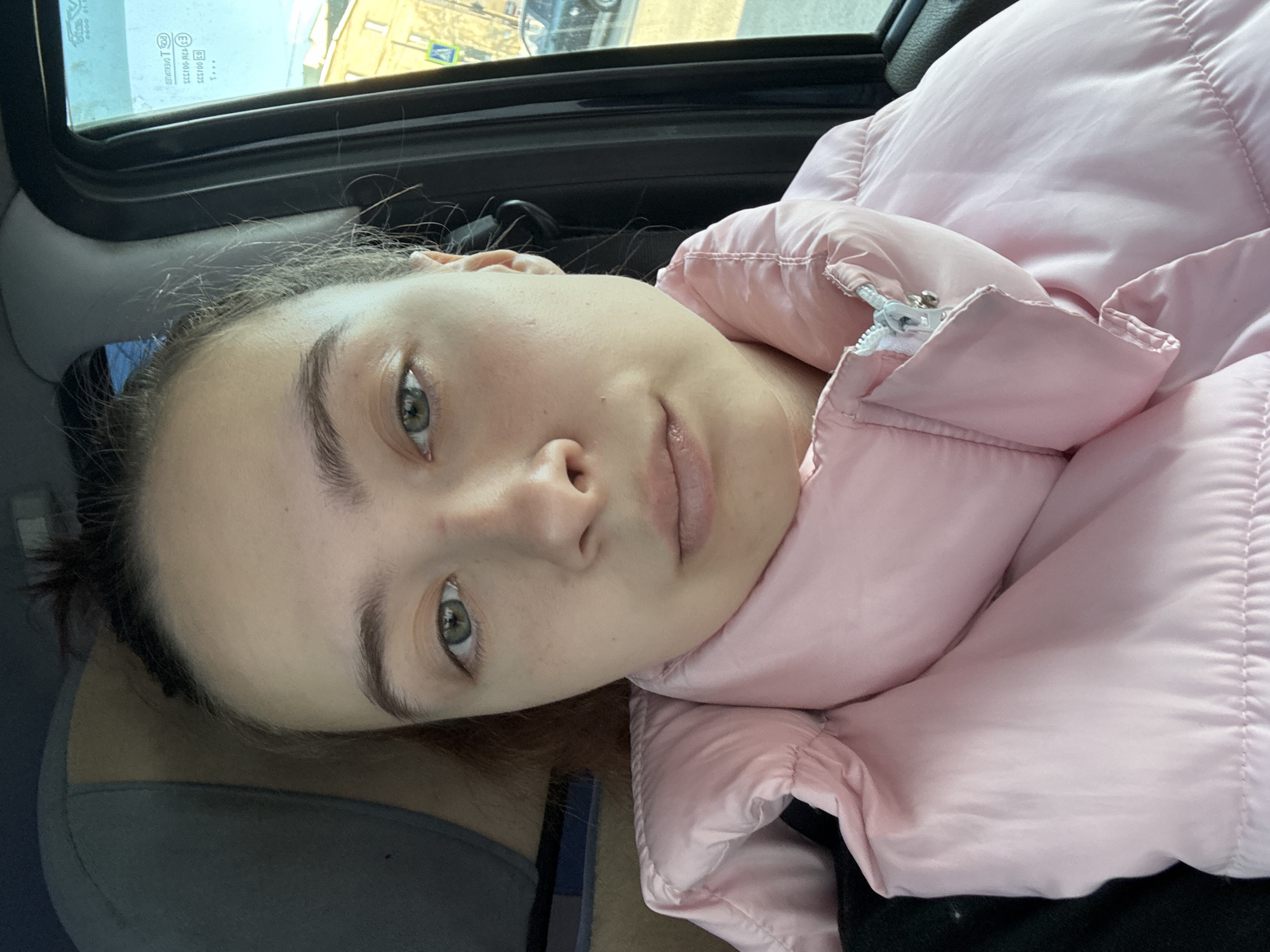 A close-up, portrait-style shot depicts a fair-skinned young woman with light eyes and dark eyebrows, wearing a light pink puffer jacket. She is reclining, possibly in a vehicle, with the car's window and roof visible above and to the side of her head. The background shows blurred buildings, suggesting an urban environment. The image does not definitively identify the location as Chaikovsky, Russia; that information is not discernible from the visual content.