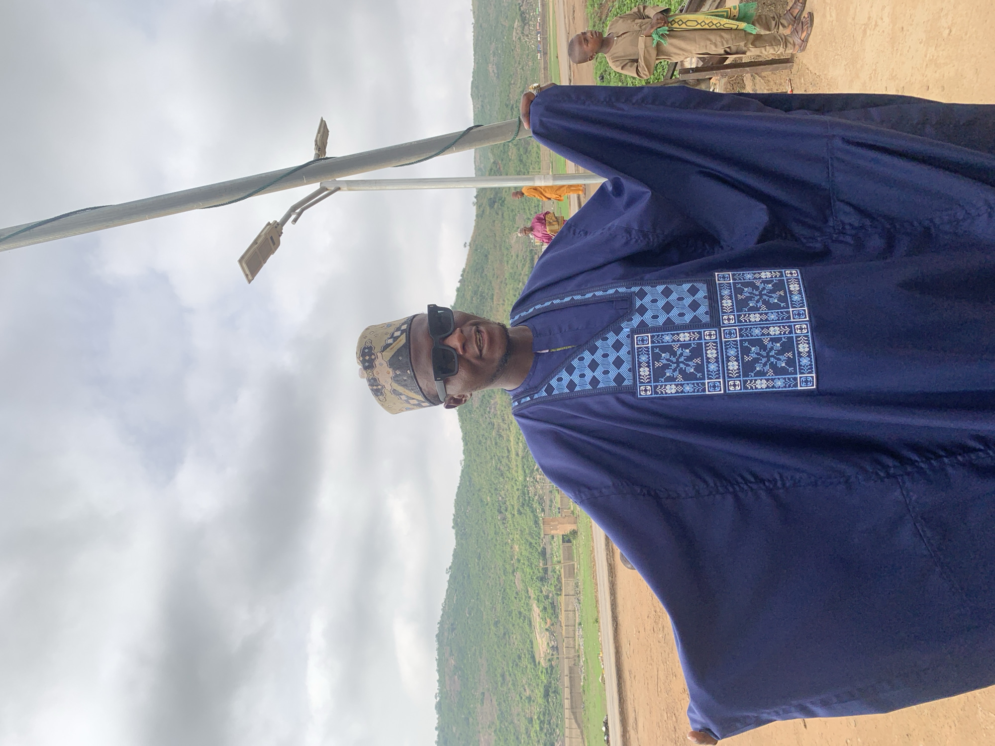 The image depicts a man wearing a dark blue traditional garment with intricate blue embroidery on the chest area and a traditional hat. He is also wearing sunglasses. The background shows a landscape with a green hill, cloudy sky, and what appears to be a street pole.
In the background, there are other people present, including a child. The location is Jos, Nigeria, as indicated by the person's attire and the environmental details.
