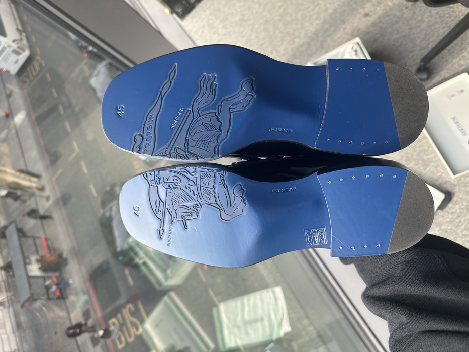 The image displays a pair of new, black leather dress shoes held with their vibrant blue soles facing the viewer, positioned above a reflective glass surface. Each shoe sole is embossed with the Burberry Equestrian Knight Device logo, the word "BURBERRY," and "PRORSUM" (on the right shoe's sole), along with the size "45" and "MADE IN ITALY." The heel sections are blue with a dark grey rubber cap.
Through the glass surface, a street scene in London, United Kingdom, is visible. The street exhibits active vehicle traffic, including a white bus on the right and a dark sedan, with road markings such as "BUS" and a reflected "STOP" sign discernible. Pedestrians are present on the pavement, and a black-framed bus stop shelter with a red roof is visible on the left, occupied by waiting individuals. A gloved hand, part of a dark garment, is holding the shoes, suggesting an individual presenting them from an elevated position within a modern building overlooking the street.