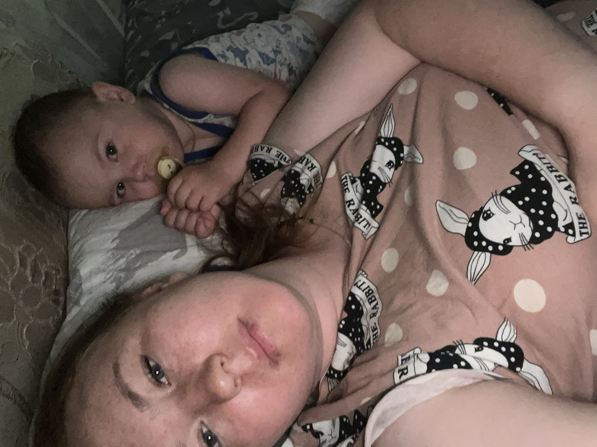 A close-up, overhead shot shows a woman and a baby lying on a bed. The woman is in the foreground, facing the camera, with her head tilted slightly. She has light skin and red hair pulled back. She is wearing a pink t-shirt with a pattern of white polka dots and cartoon rabbits. The baby is behind her, to the right, and is facing forward. The baby has fair skin and is wearing a blue and white patterned onesie. The baby has a pacifier in its mouth and is holding onto its own hands. The bed appears to be covered in patterned sheets and blankets, with a textured headboard visible at the top of the frame. The lighting is dim, suggesting an indoor setting at night or in low light conditions. The overall scene conveys a sense of intimacy and comfort, possibly a mother and child resting together. There is no information to suggest the location is in Chaikovsky, Russia.