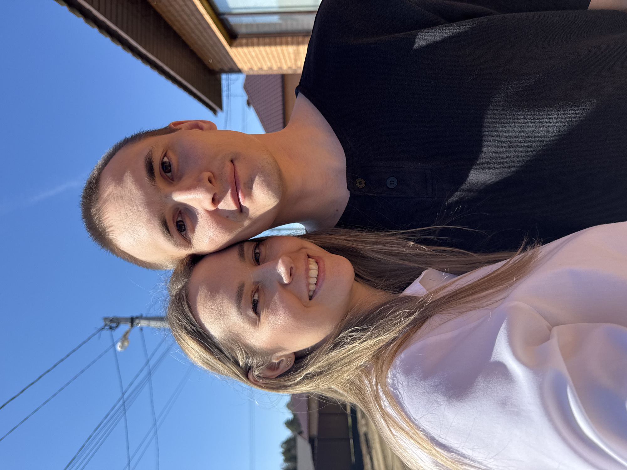 The image is a selfie of a couple outdoors. The male is on top with short hair, wearing a black shirt with buttons. The female is beneath him with long blonde hair and a white shirt. They are smiling at the camera. The background includes a clear blue sky, power lines, a building with a brown roof and brick facade, and some trees. There is no verifiable information in the image that confirms it was taken in Chaikovsky, Russia.