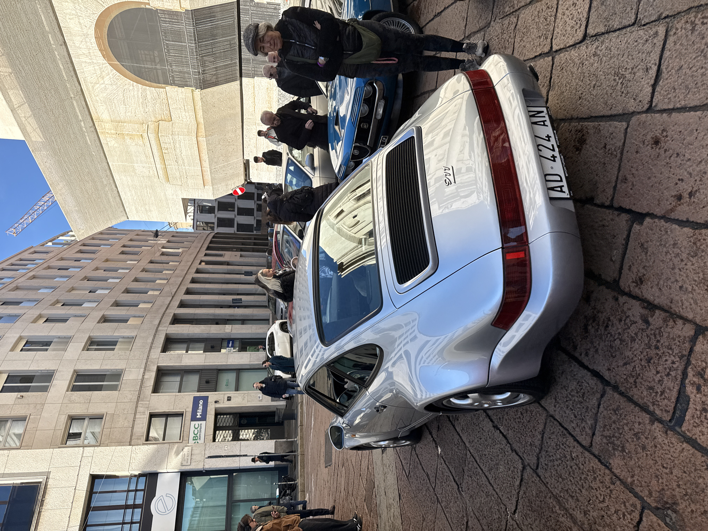 The image depicts a daytime street scene in Piazza Degli Affari, Milan, Italy, characterized by a cobblestone road and multi-story buildings. A silver Porsche 911 (likely 964 or 993 generation) is prominently parked in the foreground, facing away from the viewer, displaying Italian license plate "AD 424 AN". The car's rear features a black louvered engine cover and a continuous red taillight strip.
Behind the Porsche, a blue classic car, possibly an Alfa Romeo GTV, is partially visible, showing its front grille, headlights, and wire wheels. Various other vehicles are parked further down the street, including a white SUV.
Approximately fifteen individuals are present on the street and sidewalks. In the right mid-ground, a woman in a grey flat cap and black quilted jacket stands with her arms crossed, looking towards the camera, with a green shoulder bag slung across her. Several men are gathered near the blue classic car. On the left, near a building with a "BCC Milano Credito Cooperativo" sign, a man wearing headphones is looking at a phone, while another man in a fedora walks past.
The surrounding buildings are constructed of light-colored stone or concrete. The building on the left, housing the bank, has large rectangular windows. The building on the right features large arched windows and appears to be under construction or renovation, partially covered by scaffolding or netting, with a prominent construction crane visible against a clear blue sky above it. The street is paved with dark, irregularly shaped cobblestones, and sunlight casts distinct shadows.