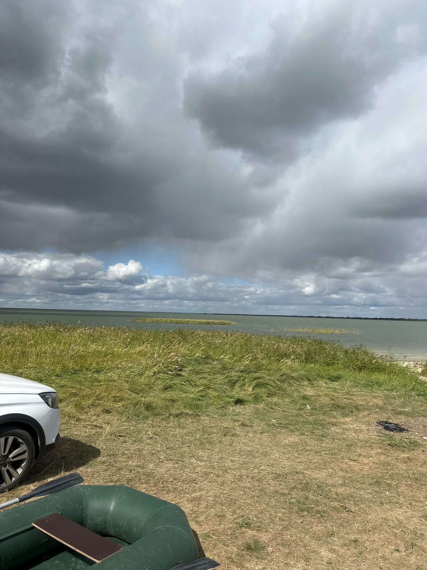 The image depicts a natural landscape with a body of water and vegetation under a cloudy sky. In the foreground, a partially visible white car is parked on dry grass. Beside the car, a green inflatable boat with wooden planks and oars is laid out. The middle ground is dominated by tall, dry grass and reeds lining the shore of a calm, green lake. The background shows the expanse of the lake meeting the horizon, with a dark, overcast sky filled with dramatic clouds. The location is identified as Tsabeleuka, Kazakhstan.