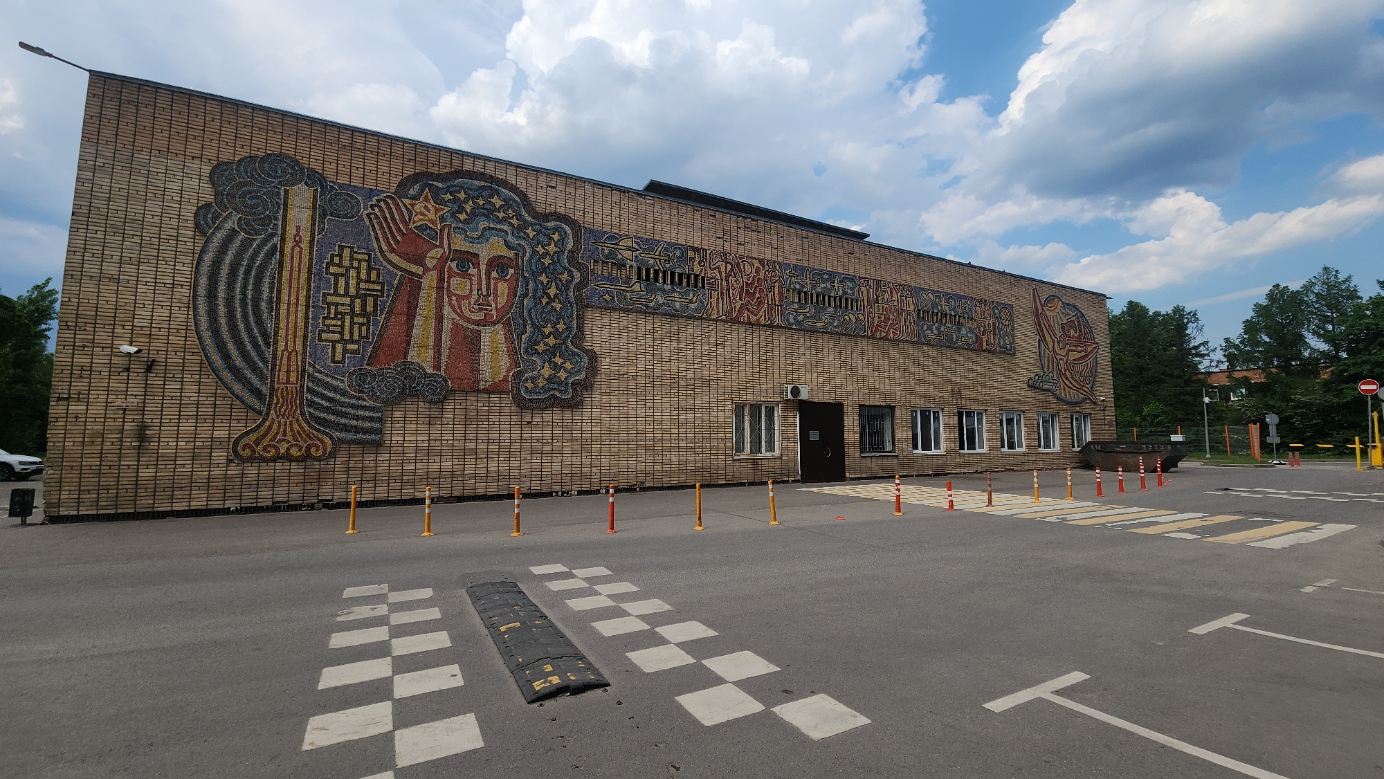 A brick building with mosaic murals is located in Khimki, Russia. The building has several windows and a door along its facade. A crosswalk with speed bumps leads up to the building. Small orange and white posts line the area in front of the building. The sky above is partly cloudy. A security camera is visible on the left side of the building. A black metal dumpster sits on the right side of the building near the entrance. A "Do Not Enter" sign is on a post further to the right.