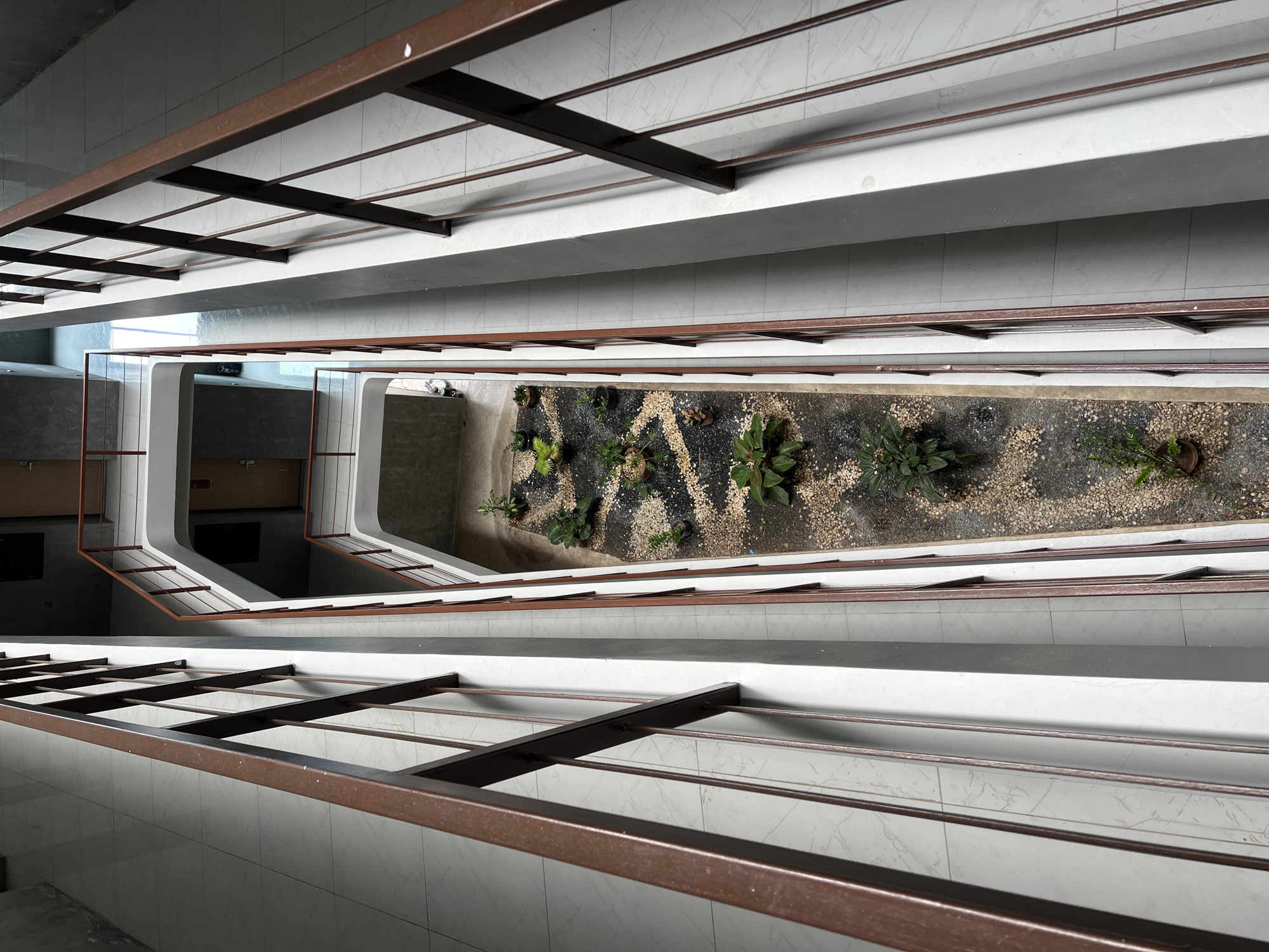 The image depicts an interior view looking down into a rectangular open space featuring plants and gravel. The perspective is from an elevated position, suggesting a multi-story building.
The foreground and background are composed of repetitive structures, likely railings or architectural supports, with a brown-colored framework and light-colored walls or paneling in between. The flooring, seen in the upper and lower portions of the frame, consists of large, light-colored tiles with faint patterns.
In the center, the rectangular open area is filled with a mix of small plants and dark gravel or pebbles. The plants appear to be a variety of small shrubs and ground cover. The area is likely a garden or landscaped feature within the building.
There are no people visible in the image. The setting seems to be modern or contemporary architecture, but specific details like city, time, or weather are not apparent.