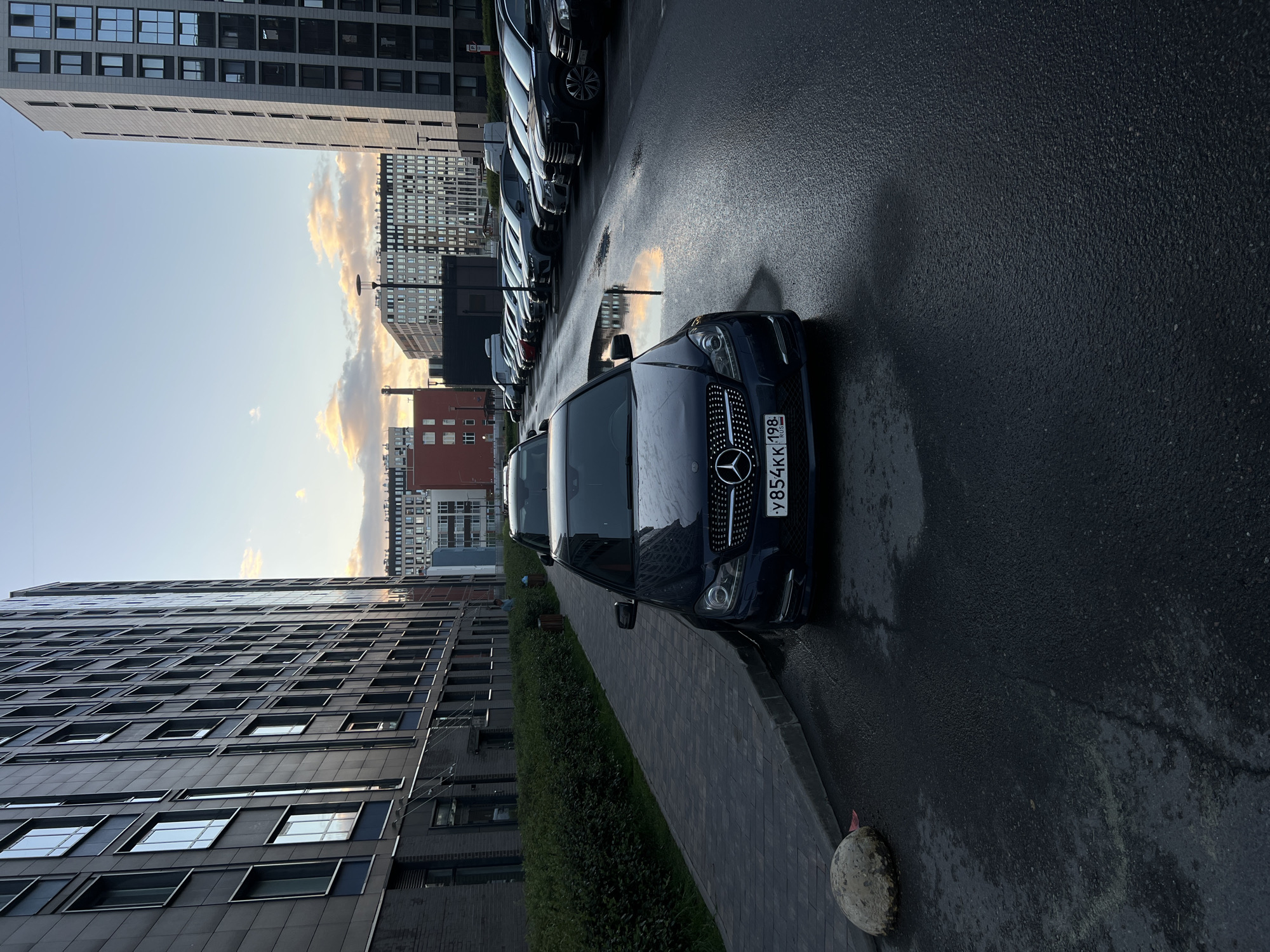 A dark blue Mercedes-Benz C-Class sedan is parked in a wet asphalt parking lot in front of a modern apartment building in Saint Petersburg, Russia. The car's license plate reads "Y854KK 198 RUS". The parking lot is lined with other cars and is reflecting the sky and the surrounding buildings. The buildings are tall and modern, with many windows. The sky is partly cloudy with hints of sunset colors. A sidewalk with a concrete bollard is visible to the left of the car. A row of dense green bushes separates the sidewalk from the building on the left. The overall scene depicts an urban environment after a rainfall.