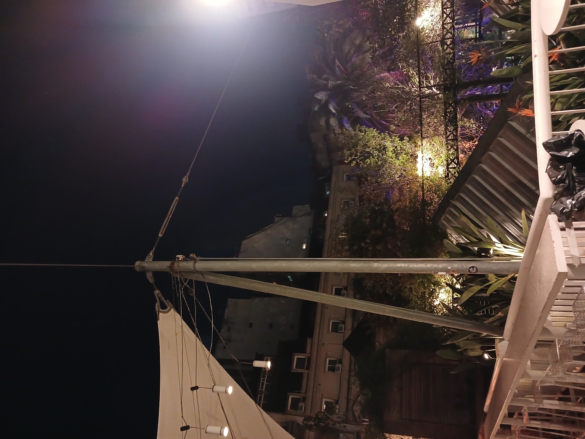At night, an unconventional upward-facing shot captures a confluence of urban and maritime elements. A portion of a white sail is visible, illuminated by integrated lights, connected to a metal mast and supporting wires. The mast is affixed with a cable that reaches to the upper left. Behind the mast and sail, a series of residential buildings with visible windows are seen. The upper right of the frame features elaborate decorative foliage, interspersed with metal lattice structures and bright accent lighting, suggesting an upscale outdoor dining area or rooftop bar. The scene is set against a dark night sky. The location is suggested to be Buenos Aires, Argentina.