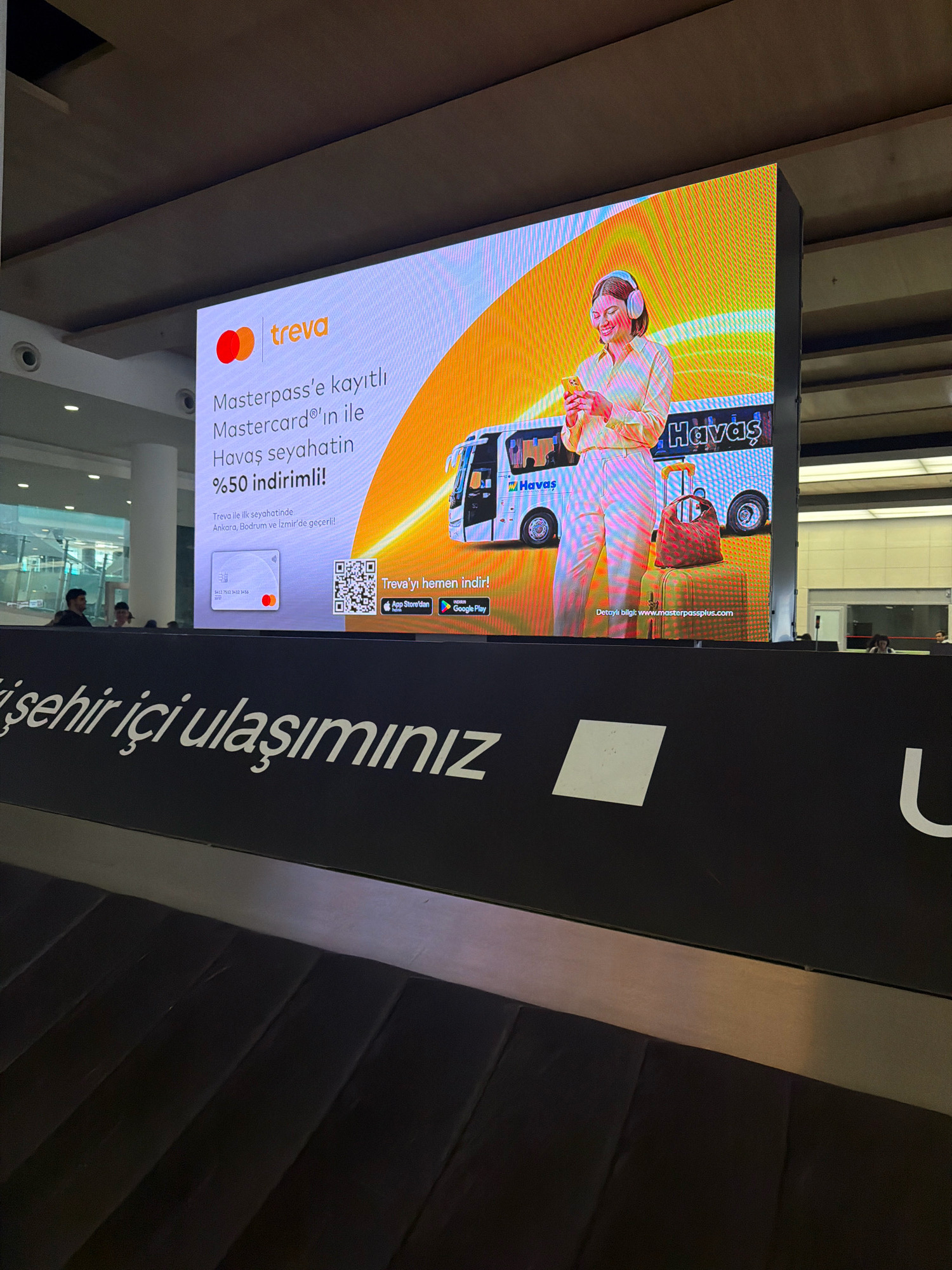 This is a photograph of a large digital billboard advertising for Treva and Havaş, likely located in Gaziemir, Turkey, given the context of Havaş being the prime ground handling services company in Turkey's airports. The billboard displays a vibrant orange and yellow background with text and images related to travel. In the foreground, a black conveyor belt with ribbed surfaces is visible, suggesting a luggage claim area at an airport.
The billboard's content promotes a discount on Havaş air travel for Mastercard users registered with Masterpass. The text on the billboard reads: "Masterpass'e kayıtlı Mastercard'ın ile Havaş seyahatin %50 indirimli!" which translates to "With your Masterpass registered Mastercard, your Havaş travel is 50% off!". Below this, it specifies that the offer is valid for the first Treva travel in Ankara, Bodrum, and Izmir. An image of a Mastercard is shown, along with QR codes for downloading the Treva app from the App Store and Google Play. A smiling woman wearing headphones is depicted holding a smartphone, and a Havaş bus and luggage are also visible in the advertisement.
The environment appears to be an indoor public space, possibly an airport terminal, with a ceiling and lighting fixtures visible in the upper part of the image. The lower portion of the image shows the black conveyor belt, and the text "şehir içi ulaşımınız" which translates to "your urban transportation", suggesting a broader service offering beyond air travel. Several individuals are partially visible in the background, appearing to be passengers or staff, but their details are indistinct. The overall scene captures a promotional display in a transportation hub.