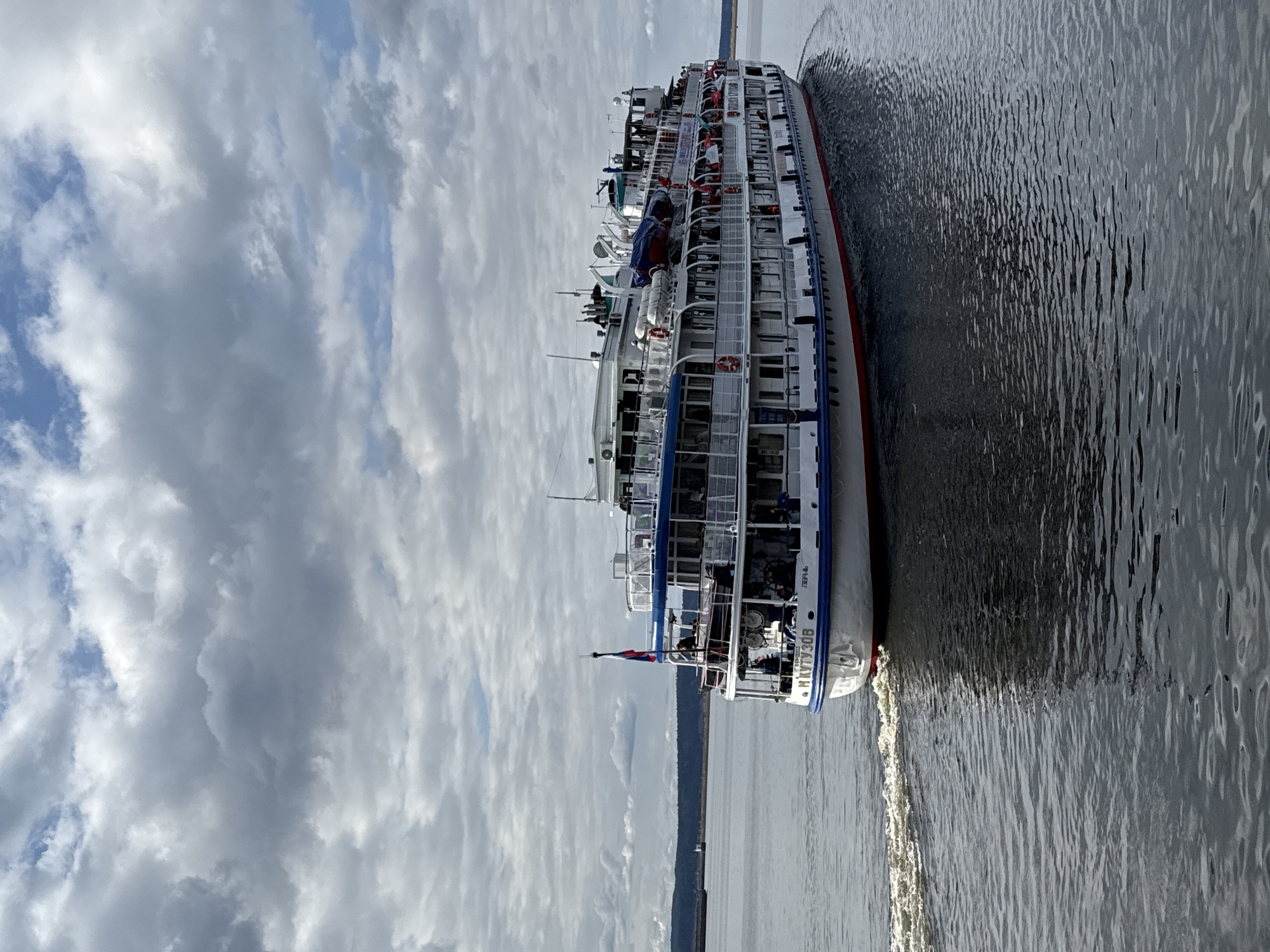 A large, multi-deck passenger ship is seen cruising on a wide body of water under a partly cloudy sky. The photograph is taken from a slightly elevated perspective, showing the starboard side of the ship as it moves, leaving a visible white wake behind it.
The ship is predominantly white, featuring blue trim along its decks and a distinctive red stripe along its hull near the waterline. It possesses multiple levels with numerous windows, balconies, and open deck areas. Several passengers are visible on the upper and mid-decks, enjoying the journey. Life preservers (orange rings) and blue-covered lifeboats are secured on the upper decks. Various antennas and radar equipment are mounted on the ship's roof. A small Russian flag is proudly displayed at the stern.
The water surface is dark and shows ripples, reflecting the cloudy sky. In the distant background, a low-lying, dark landmass or shoreline stretches across the horizon. The sky is overcast with a mix of light and dark grey clouds, but patches of blue sky are visible, suggesting it is daytime with diffused sunlight.
The ship is actively engaged in travel, likely on a river cruise or transport. The scene appears calm and serene.
Visible text on the stern of the vessel clearly identifies it as "М. КУТУЗОВ" (M. Kutuzov), with "ПЕРМЬ" (Perm) written below, indicating its home port or registration city. A smaller alphanumeric string, possibly "707И" or "7074", is also visible. Given the location context of Chaikovsky, Russia, these details strongly suggest the ship is operating on the Kama River or an interconnected waterway.