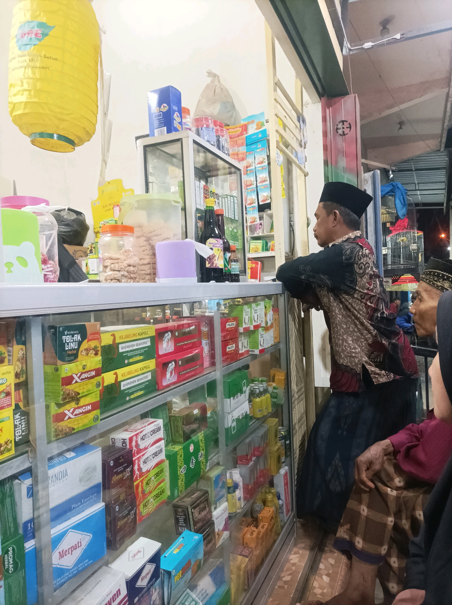 The image depicts a street-side shop in Sumberagung, Indonesia. A man wearing a black peci cap and a patterned traditional shirt is leaning on a glass display counter filled with various packaged goods, likely medicine or toiletries. Another older man, also wearing a peci cap and a maroon shirt, is partially visible on the right, looking towards the counter. Behind the counter, shelves are stocked with boxes and bottles. The shop appears to be open to the street, with a yellow lantern hanging on the left. The overall impression is that of a small, local convenience store or pharmacy.