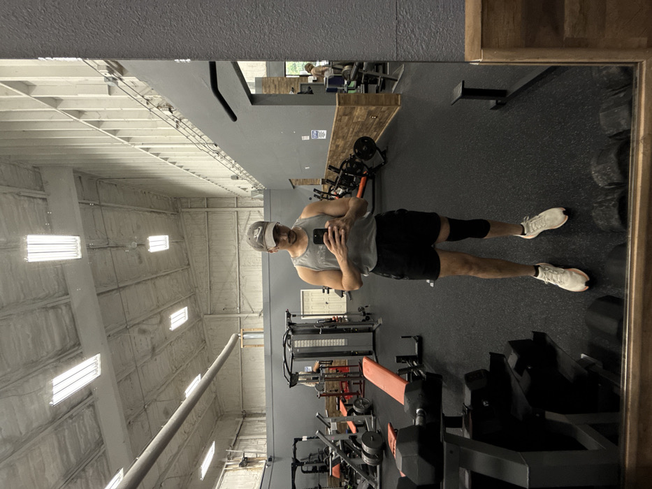 The image depicts the interior of a gymnasium in Osage Beach, United States. A male individual stands in the foreground, facing a large mirror and holding a smartphone, appearing to capture his reflection. He wears a grey baseball cap with a Nike logo, a grey sleeveless tank top, black athletic shorts, and white athletic shoes. A black knee brace is visible on his right knee. His reflection shows a mustache. The gym floor is covered with black rubber matting. Various pieces of exercise equipment are visible in the background, including a dumbbell rack with multiple black dumbbells, an orange-padded weight bench, and several multi-station weight machines. The gym features high, vaulted ceilings with exposed structural elements and white spray foam insulation. Rectangular fluorescent light fixtures illuminate the space. Walls are segmented horizontally with dark grey on the lower section, a wooden panel mid-section, and a lighter grey or off-white upper section. In the far right background, partially obscured by the mirror's frame, another individual with blonde hair is reflected, operating gym equipment.