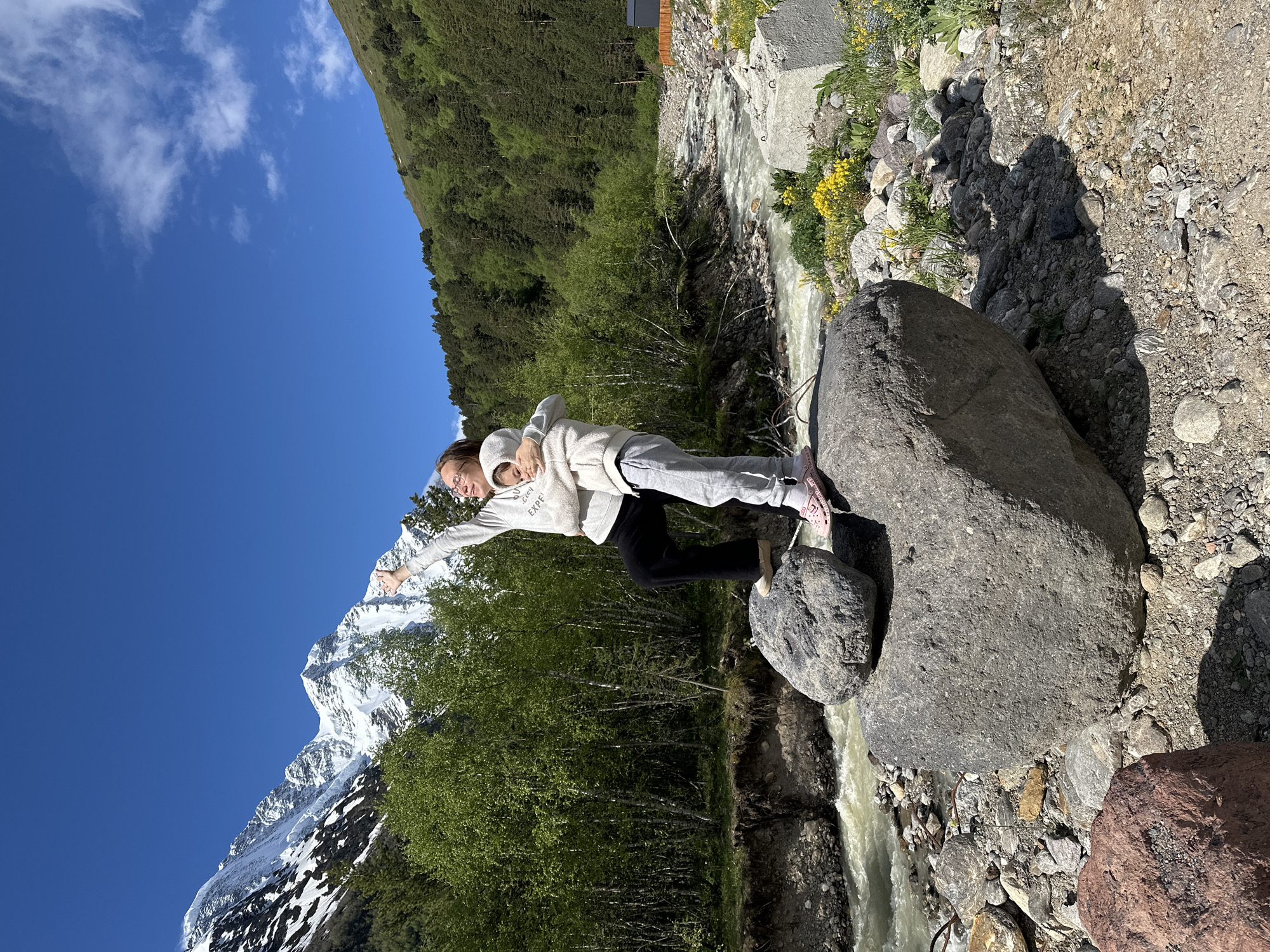 In Terskol, Russia, two individuals, a woman and a child, stand on large boulders near a river. The woman, on the left, wears glasses, a light-colored hooded sweatshirt, dark pants, and light pink sandals. She is reaching her right arm upwards and to the left. The child, on the right, embraces the woman, wearing a similar light-colored hooded sweatshirt and grey sweatpants with pink sandals. They are positioned in front of a backdrop of lush green trees, a flowing river, and snow-capped mountains under a bright blue sky with scattered clouds. The riverbank is rocky, with smaller stones and patches of yellow wildflowers visible. The overall scene captures a moment of enjoyment and connection in a mountainous natural environment.