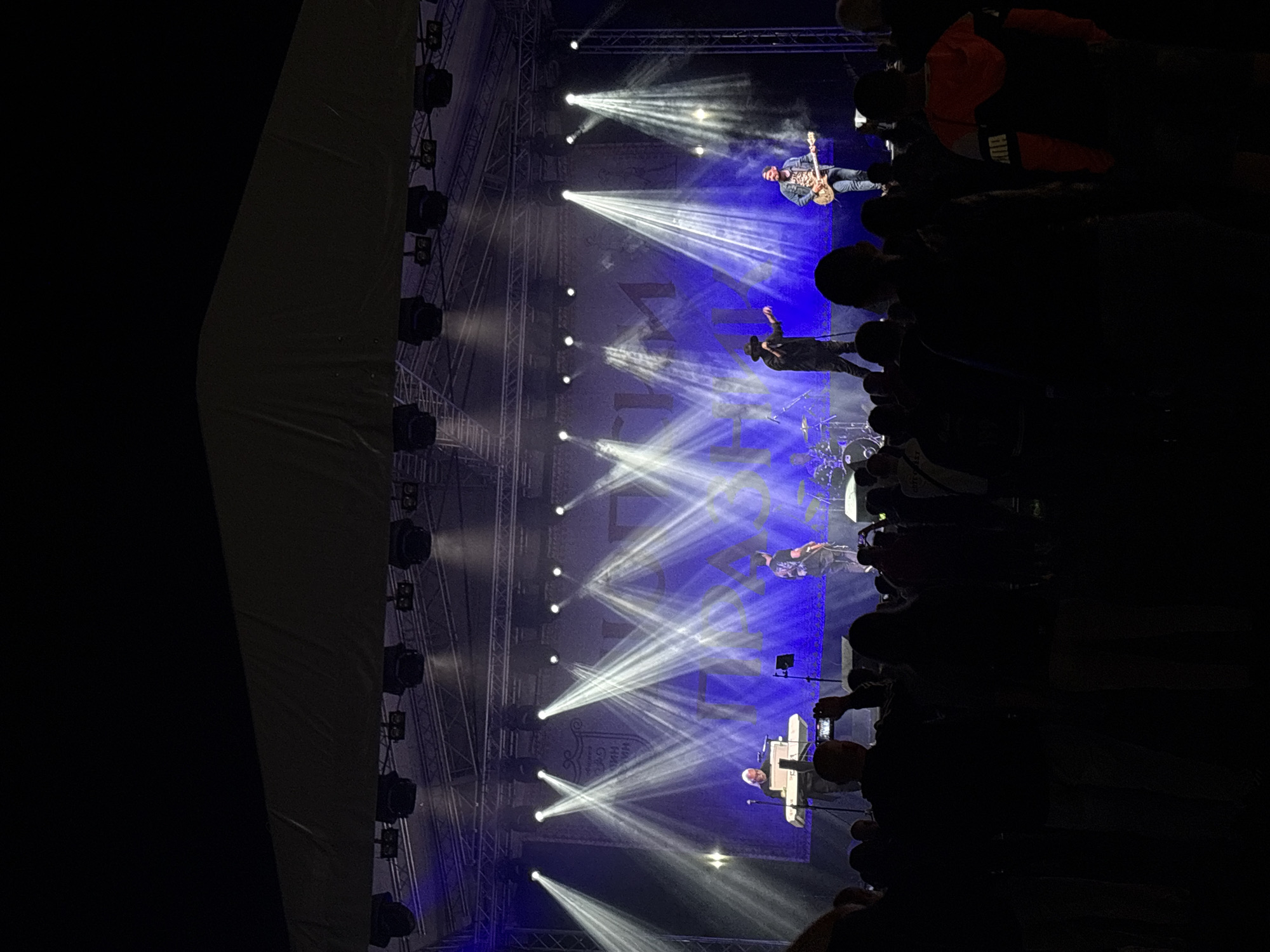 This image depicts a nighttime outdoor music concert in Elin Pelin, Bulgaria. A band is performing on a large stage under a dark canopy supported by metal trussing. The stage backdrop is a blue banner featuring stylized white text, possibly "ЕЛИН ПЕЛИН", and a crest emblem on the left. Bright white spotlights emit distinct beams of light across the stage, which is also illuminated by blue ambient lighting.
From left to right on stage, performers include:
1. A male individual with light-colored hair, wearing a dark top, seated and playing a white keyboard.
2. A male vocalist, wearing a dark hat and dark clothing, standing centrally, holding a microphone, with his right arm raised.
3. A male guitarist, wearing a denim jacket, patterned shirt, and jeans, standing on the right side of the stage, playing an electric guitar.
4. A drum kit is partially visible behind the central performers.
In the foreground and midground, a large crowd of people, mostly silhouetted against the stage lights, faces the performers. Several individuals are holding up smartphones, presumably recording the event. One person in the right foreground wears an orange and black hooded jacket. The upper portion of the image shows the dark night sky above the stage structure.