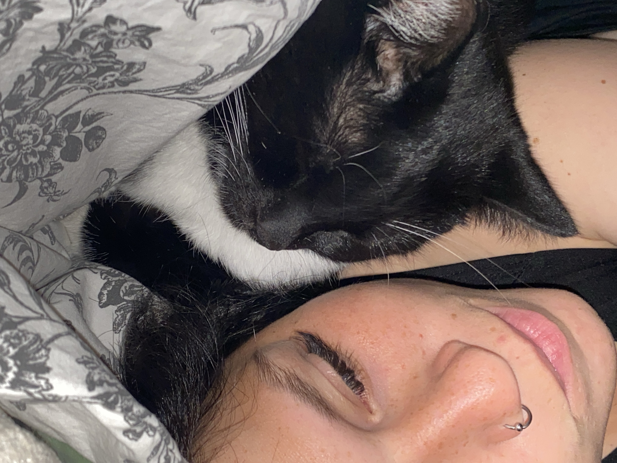The image shows a close-up view of a person and a black and white cat sleeping together. The person's face is visible on the right side of the frame, with their eyes closed and their nose pierced with a hoop earring. Their skin has freckles. The cat is nestled against the person's neck and shoulder, with its eyes closed and its fur appearing soft. They are both lying on a patterned comforter or blanket. The lighting suggests it is nighttime or indoors. The image does not contain any information or context that would allow for the determination of a location in Chaikovsky, Russia.
