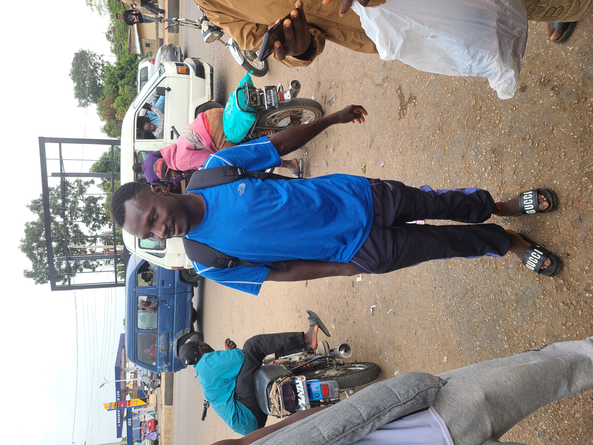 The image shows a street scene in Gura Top, Nigeria. A man in a blue shirt and black pants stands on a paved surface, with several people around him. One person in a brown jacket holds a mobile phone and a white plastic bag. Nearby, there are two motorcycles, one with a man in a blue shirt sitting on it. Two vans are parked on the side of the road, one white and one blue. A gas station is visible in the background, identified by "GASCO" signage.