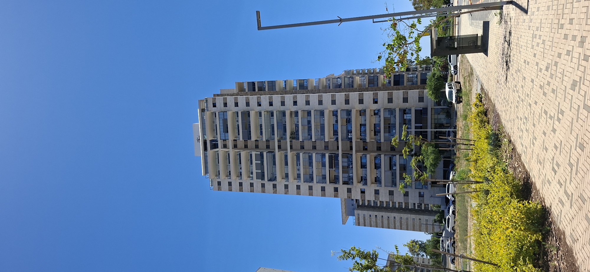 An outdoor, eye-level, full shot captures a modern high-rise apartment building against a clear blue sky. The building features a light-colored facade with numerous balconies, many enclosed with glass railings. A smaller, similar-style building is visible to the left of the main structure. In the foreground, a patterned brick walkway runs alongside a strip of vibrant green and yellow bushes. Several trees, some with sparse foliage, are interspersed along the walkway. Cars are parked on a street to the left and right of the buildings, and a modern streetlamp stands on the right side of the frame. A dark, rectangular shelter with a slanted roof is situated on the sidewalk near the streetlamp. The overall scene suggests a well-maintained urban environment on a sunny day.